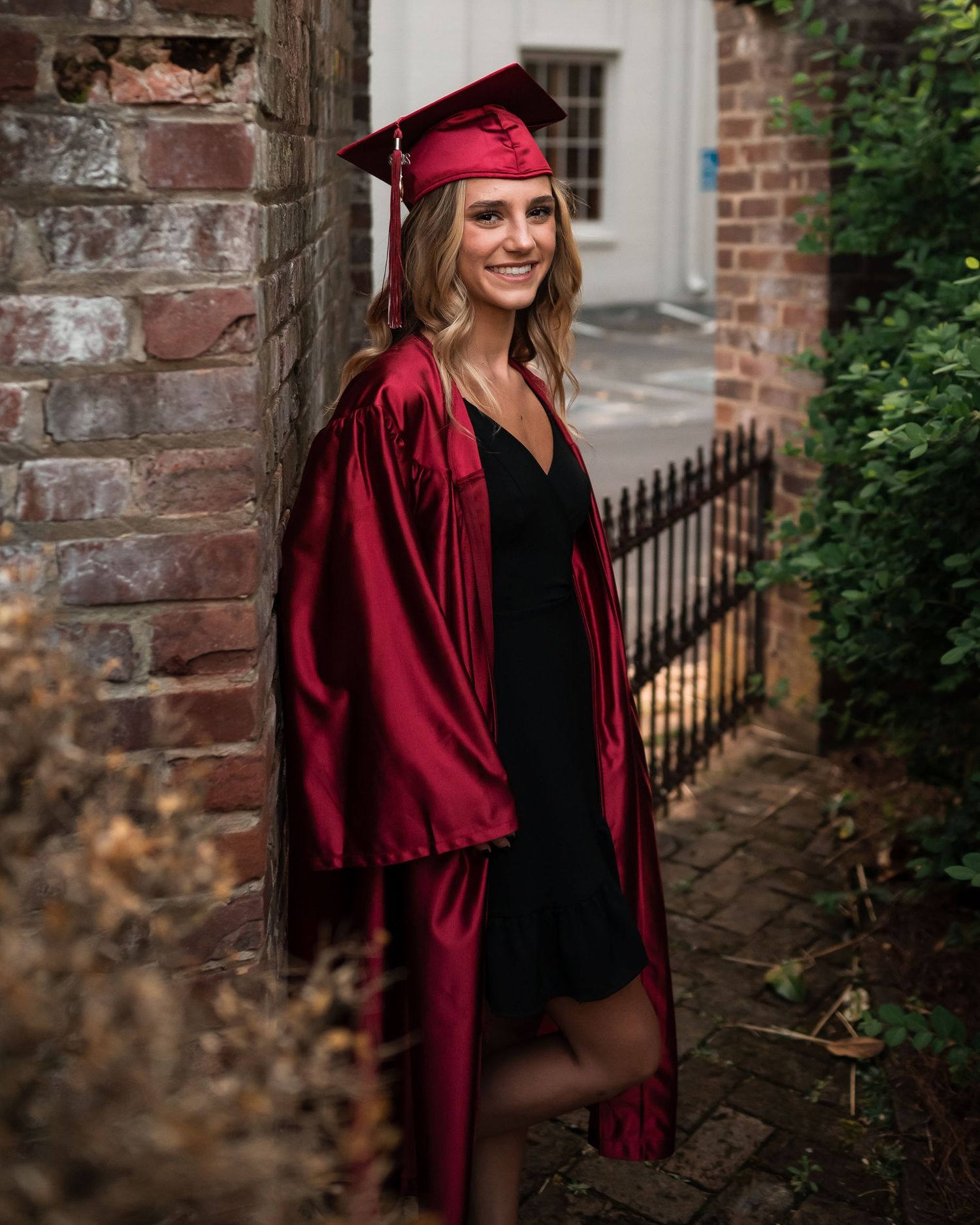 Graduate poses by brick wall in crimson cap and gown during graduation photoshoot.
