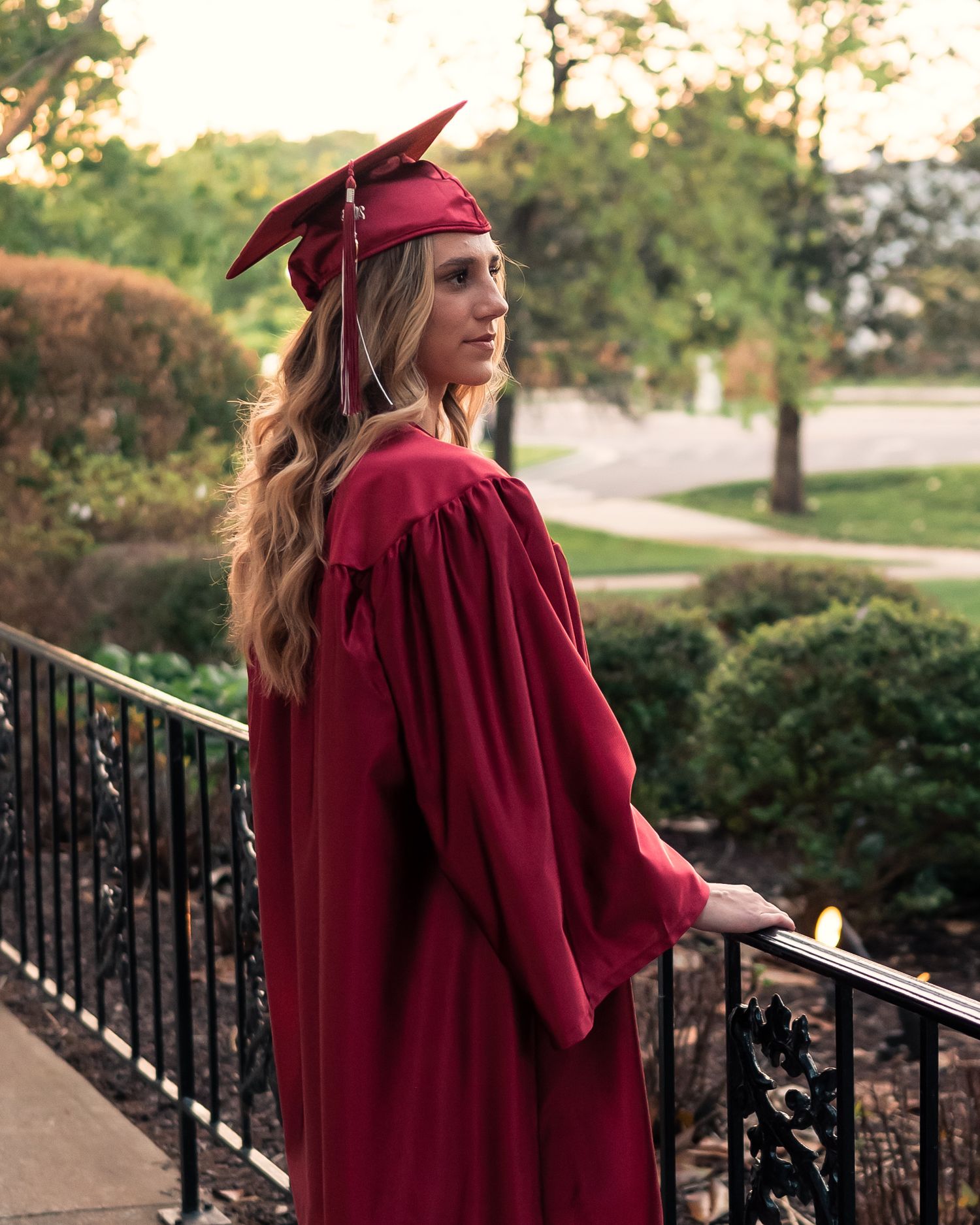 Graduate in red cap and gown poses outdoors during sunset for graduation photos.