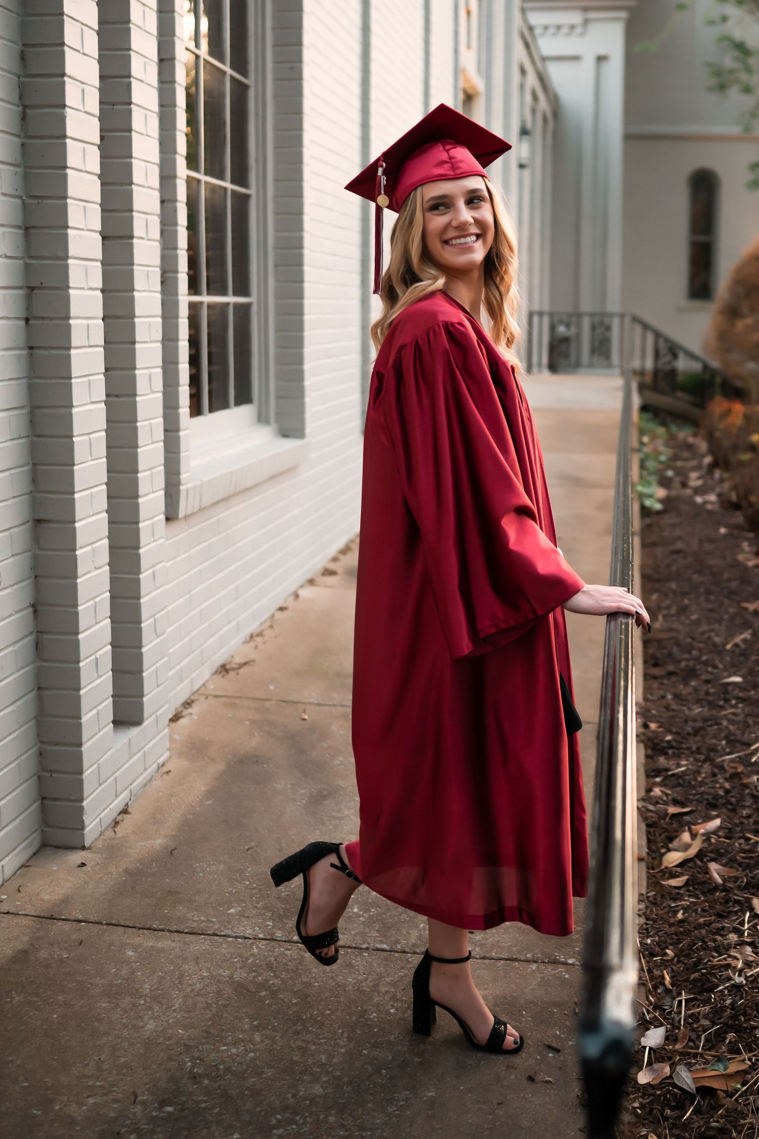 Graduate in red graduation attire poses near white columns during photo session.