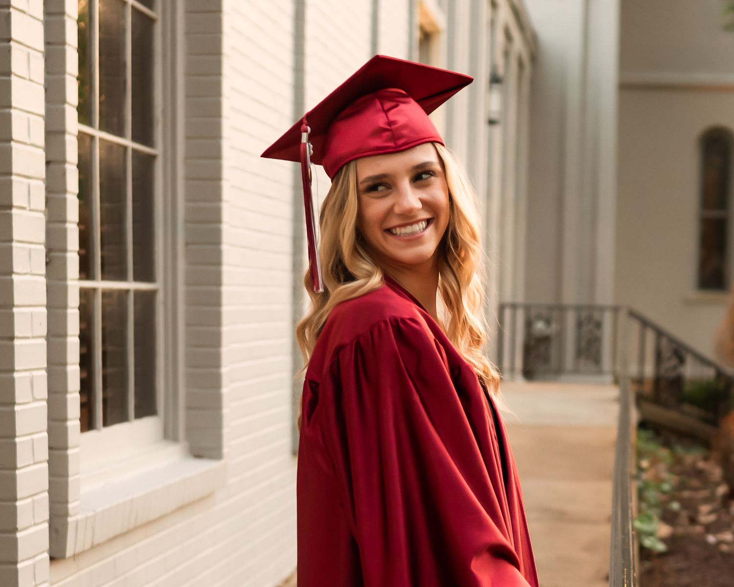 Graduate in crimson academic regalia stands in front of white brick building.