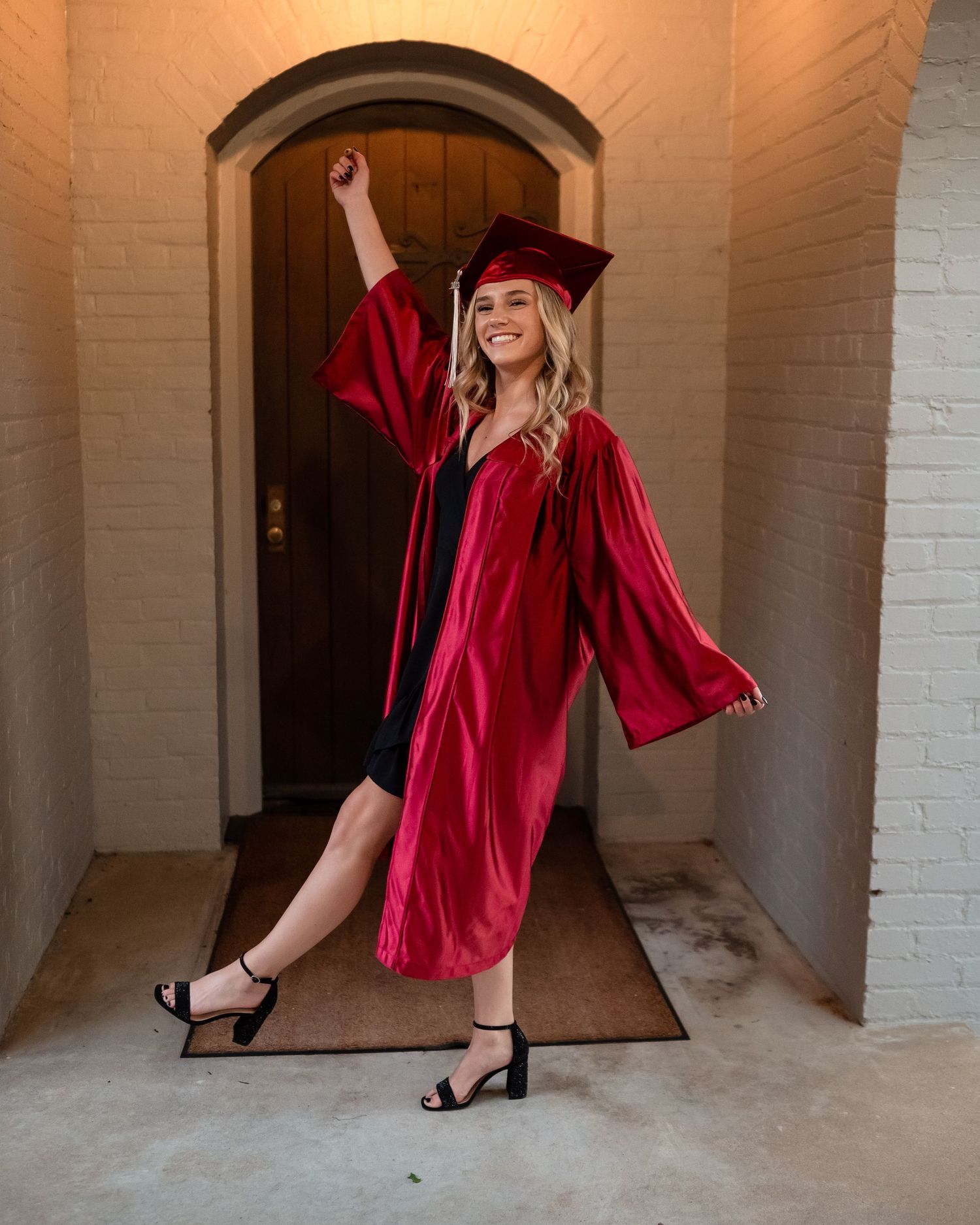 Graduate in red cap and gown poses in front of arched doorway.