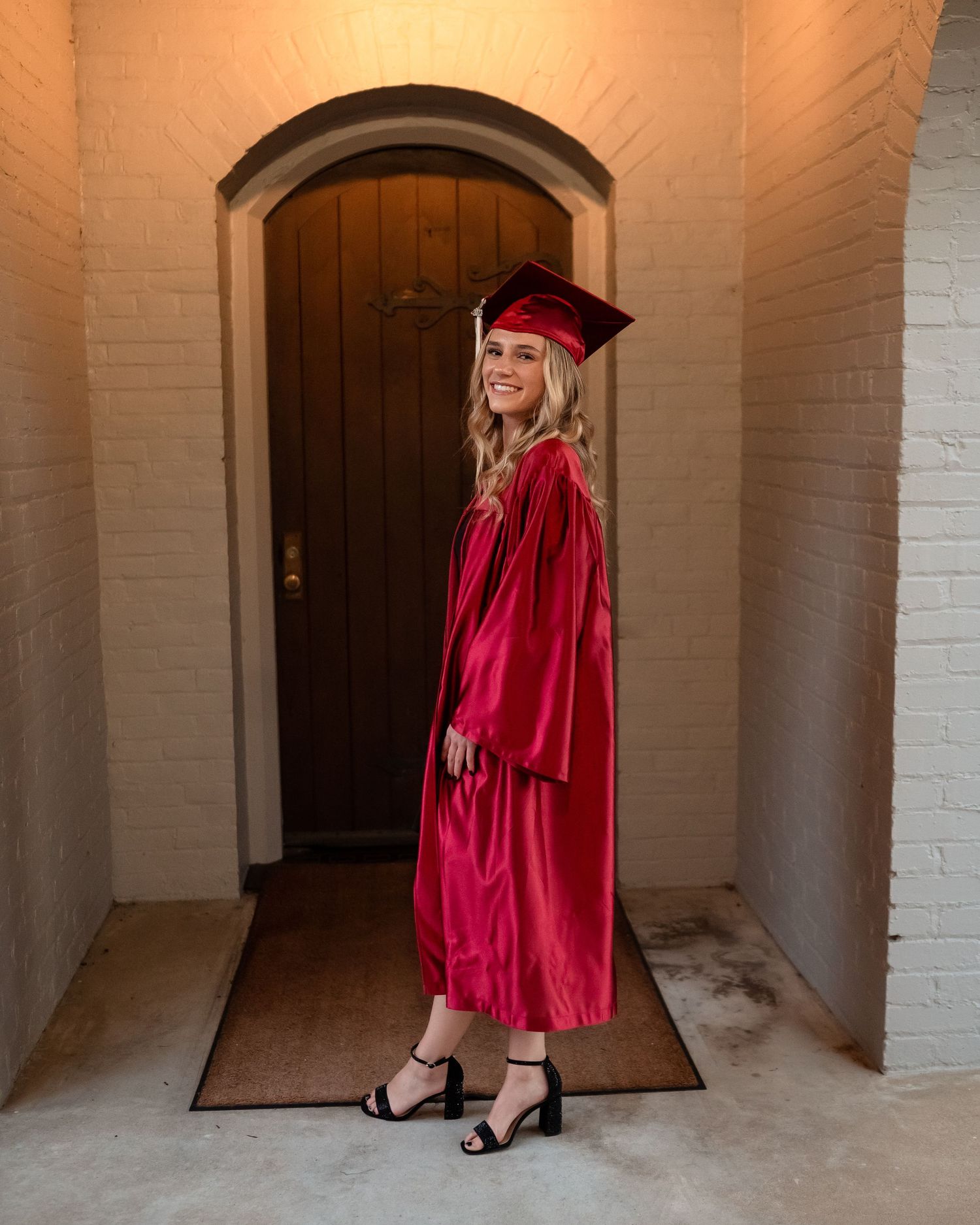 Graduate in red regalia takes photos in front of white brick archway.