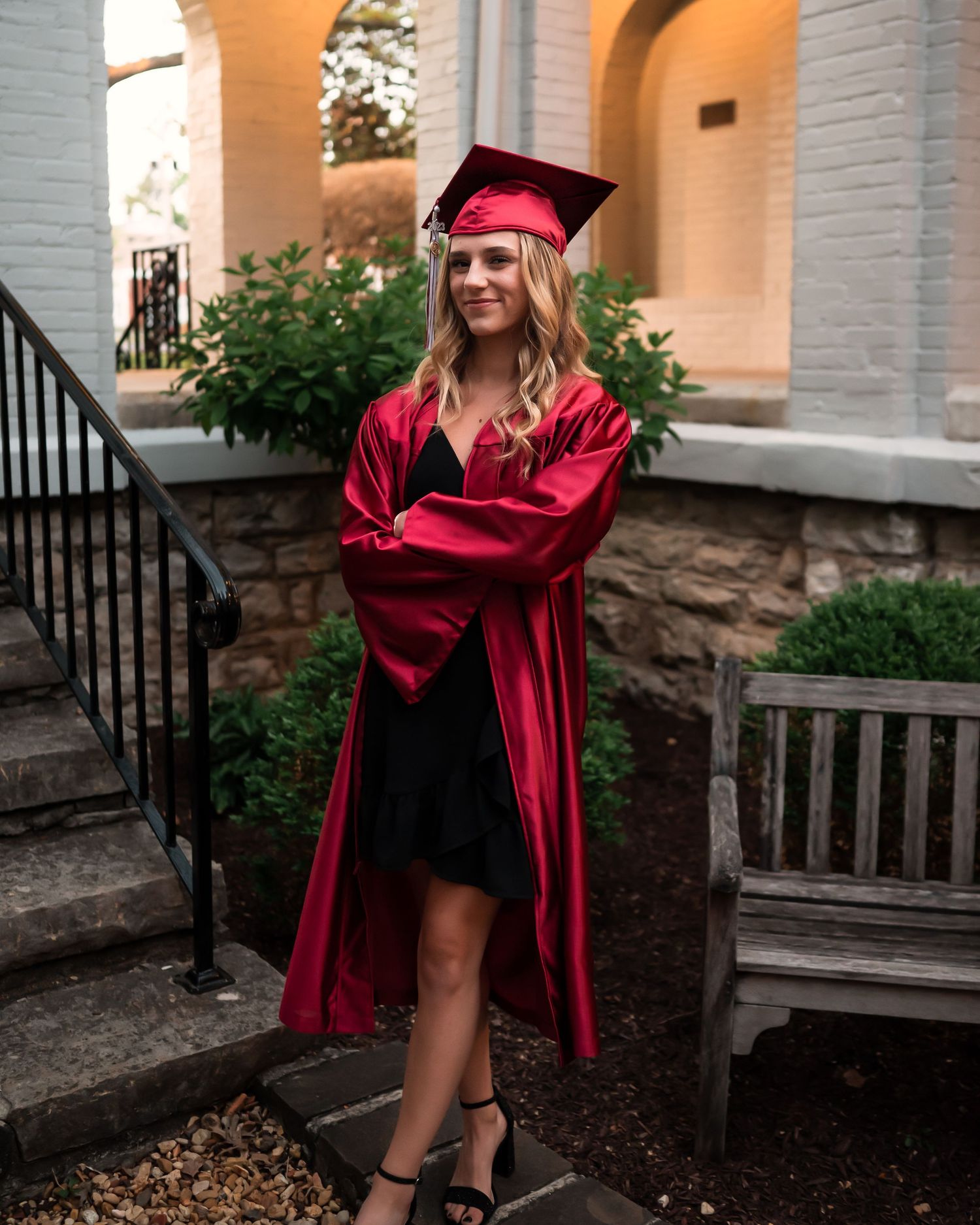 Graduate in red cap and gown poses on stone steps during outdoor photo session.