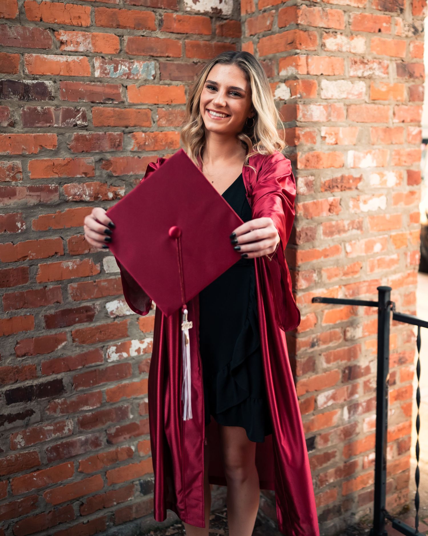 Graduate displays red graduation cap while wearing black dress and red gown by brick wall.