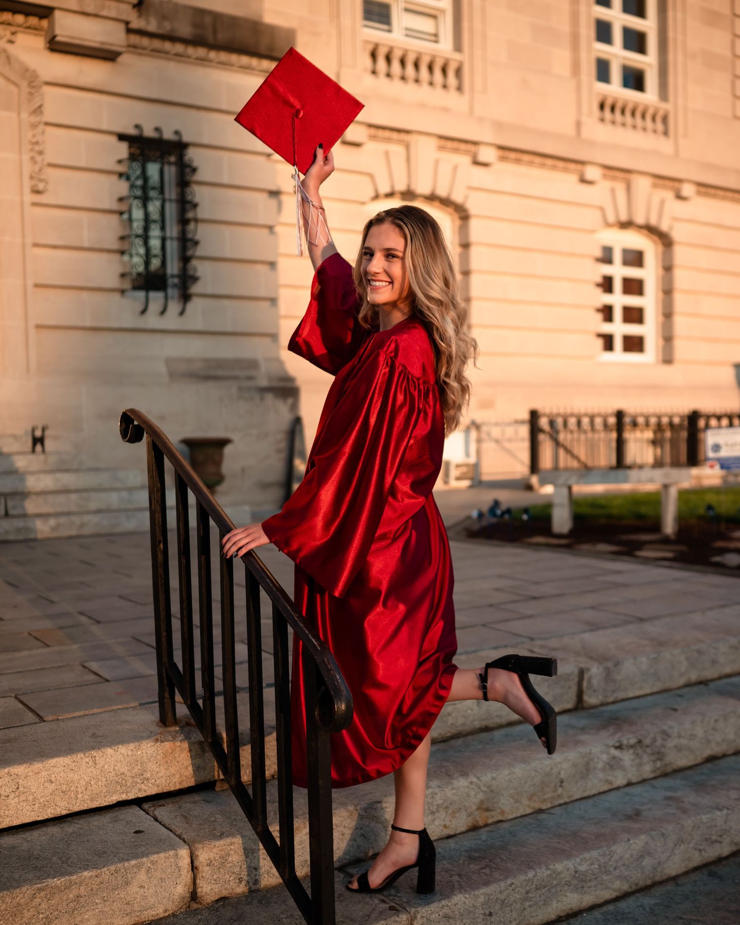 Graduate celebrates on steps holding red cap while wearing crimson graduation gown.