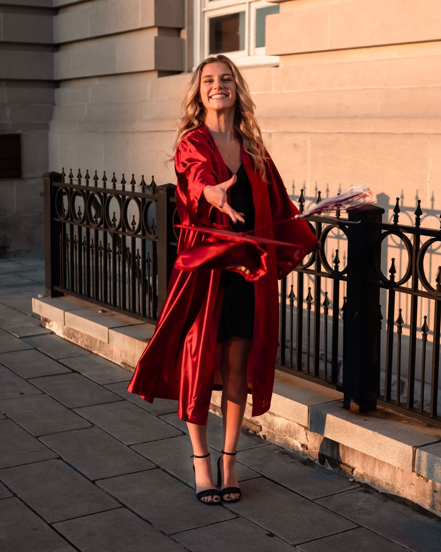 Graduate in flowing red graduation gown poses by ornate iron railing during sunset.