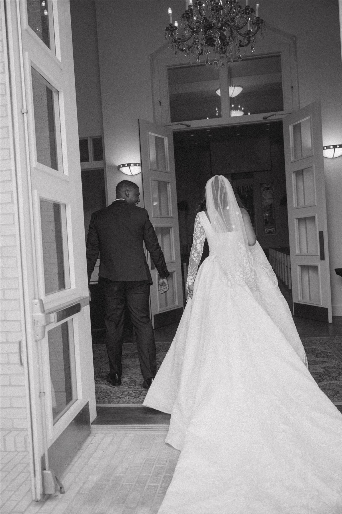 A bride in a flowing white gown and veil walks through elegant double doors with her companion at a wedding venue.