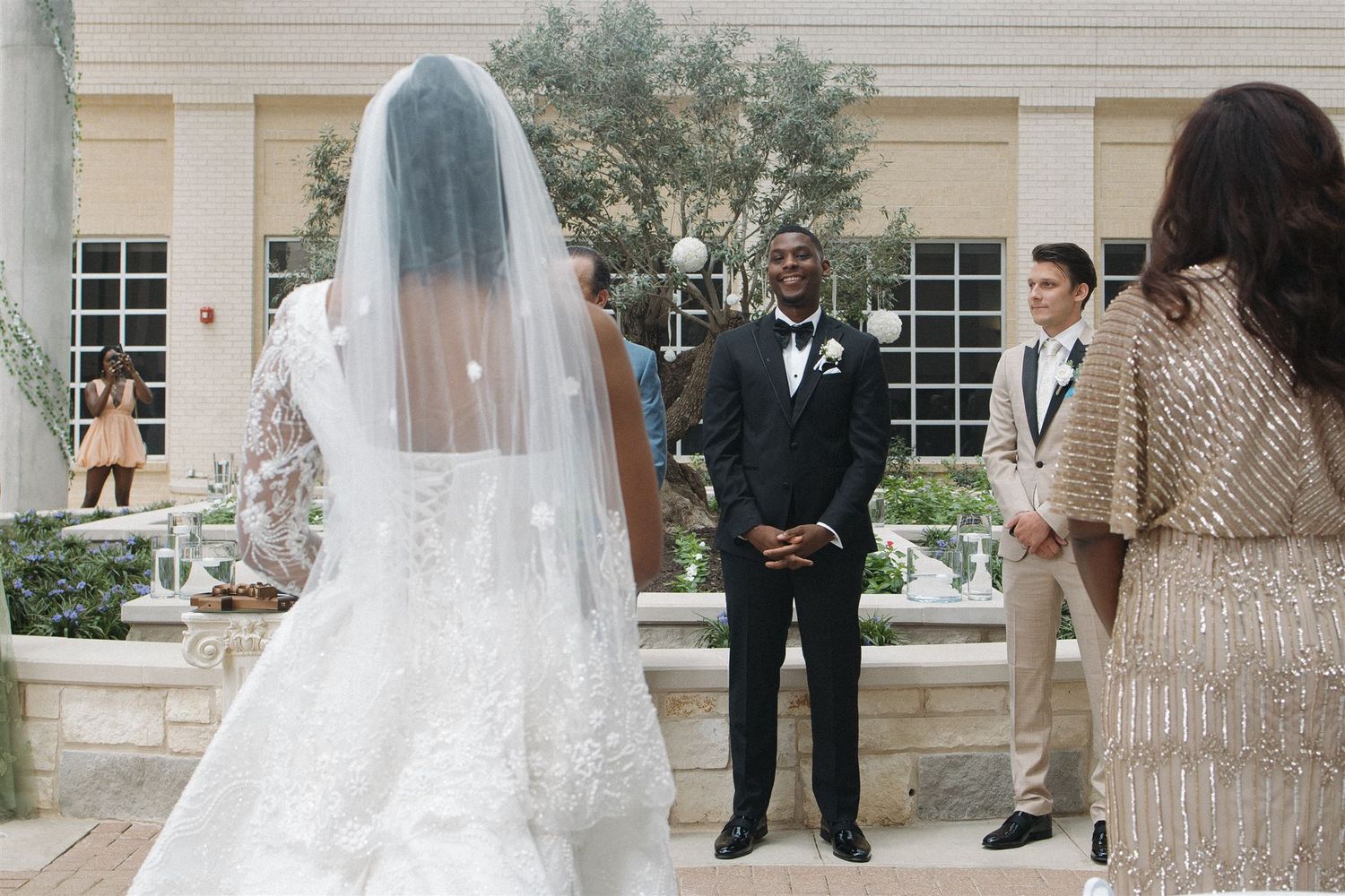 An outdoor wedding ceremony in front of a classical building with a bride in a long veil approaching the altar area.