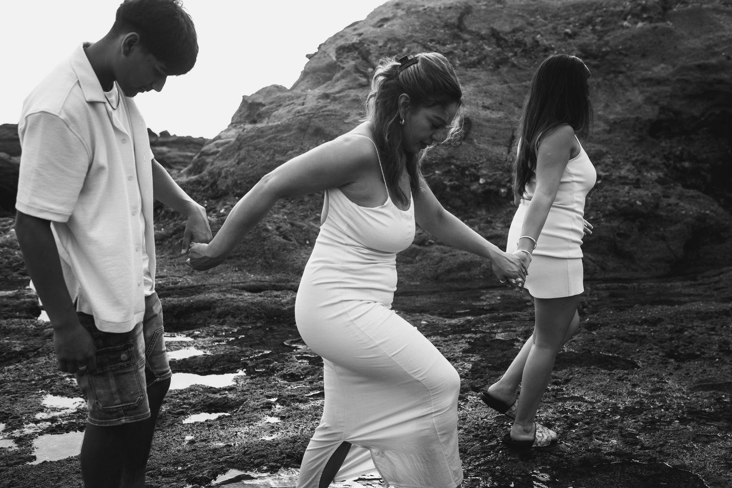 Three people walking along rocky coastal cliffs wearing white dresses.
