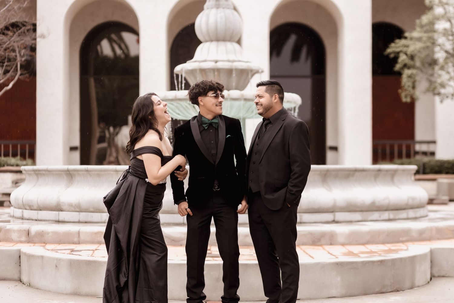 Three people in formal attire posing together against an ornate stone building facade.