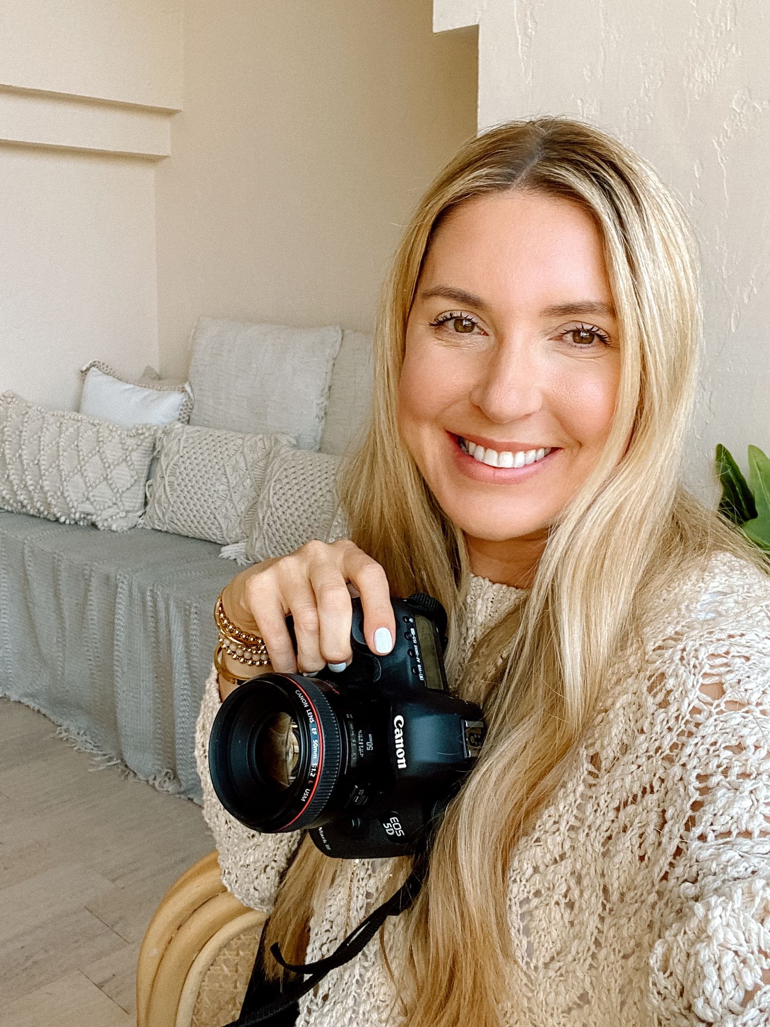 Woman in lace sweater holding professional camera and smiling in bright home setting.