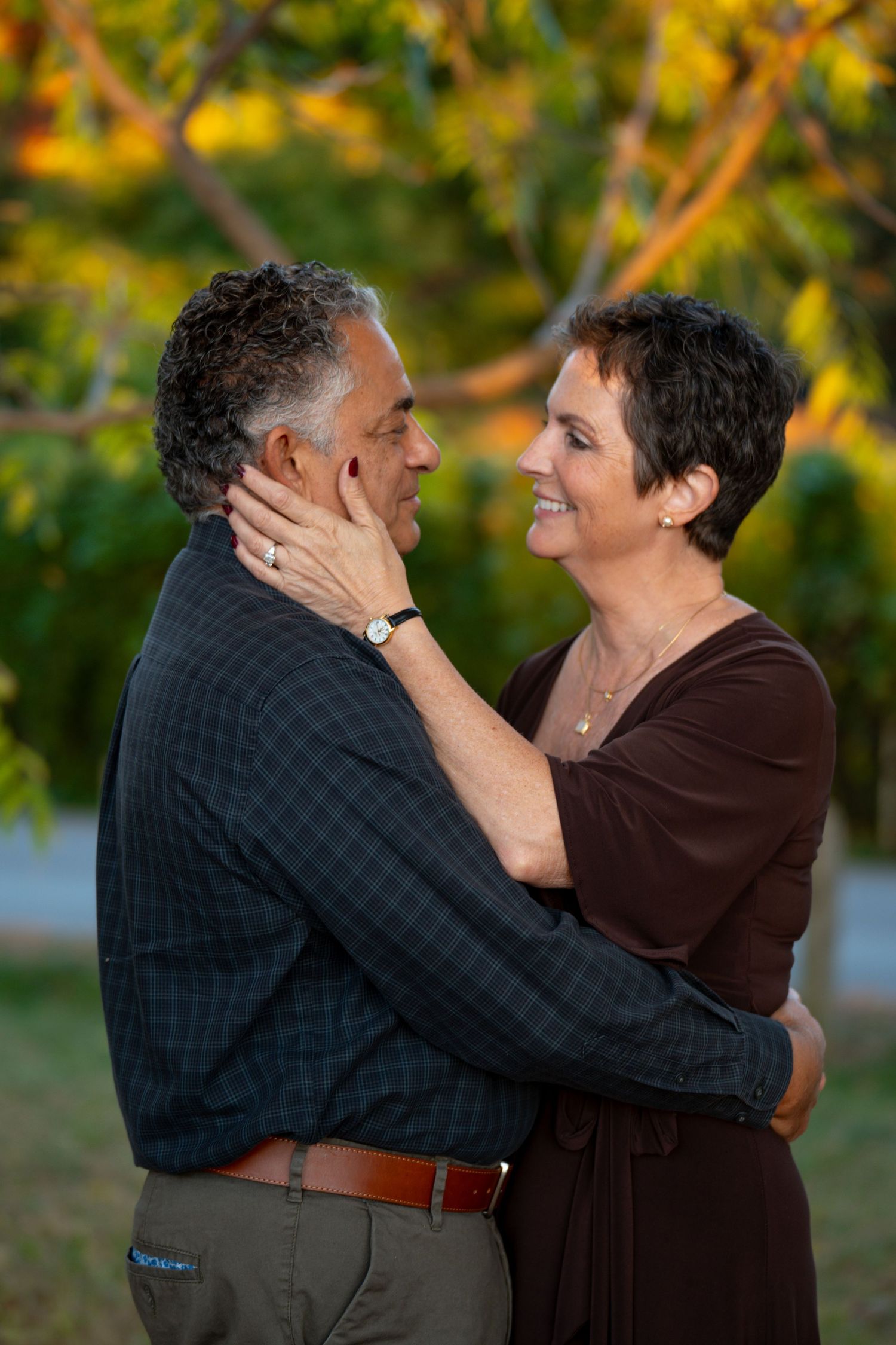 A couple shares a tender embrace outdoors with autumn foliage in the background during golden hour lighting.