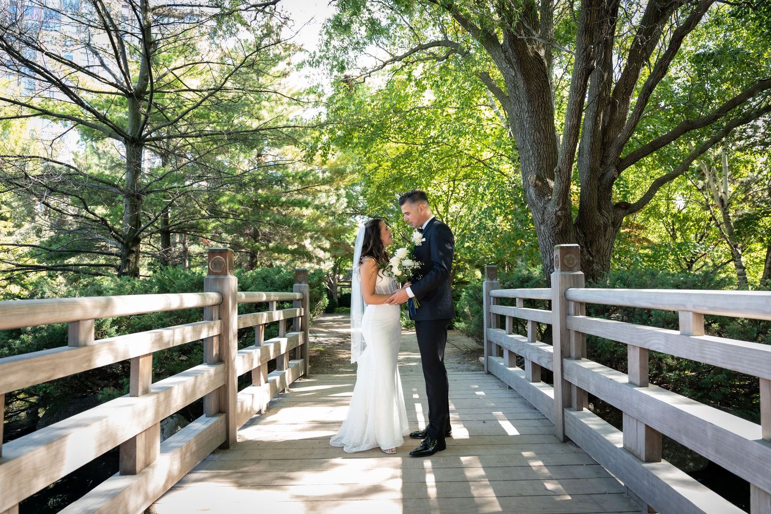 A couple walks along a scenic wooden boardwalk path surrounded by lush green trees in a peaceful outdoor setting.