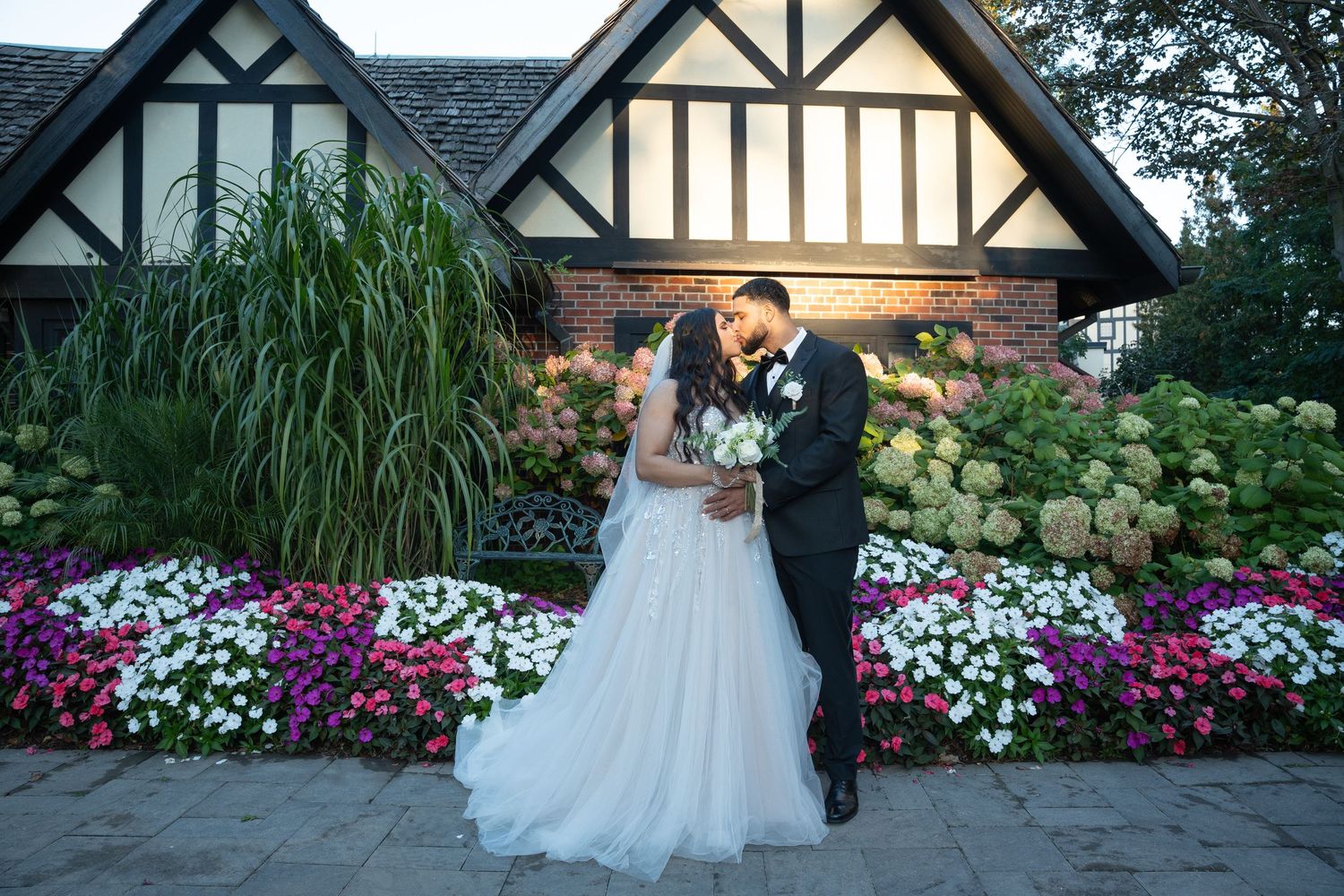A couple embraces in front of a Tudor-style building surrounded by colorful flower gardens and ornamental grasses.
