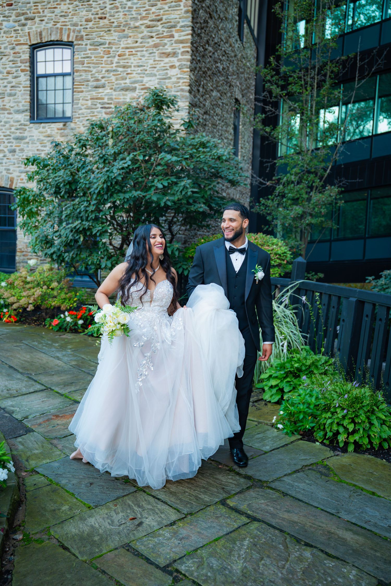 Couple in formal wedding attire sits together on stone steps surrounded by lush garden greenery.