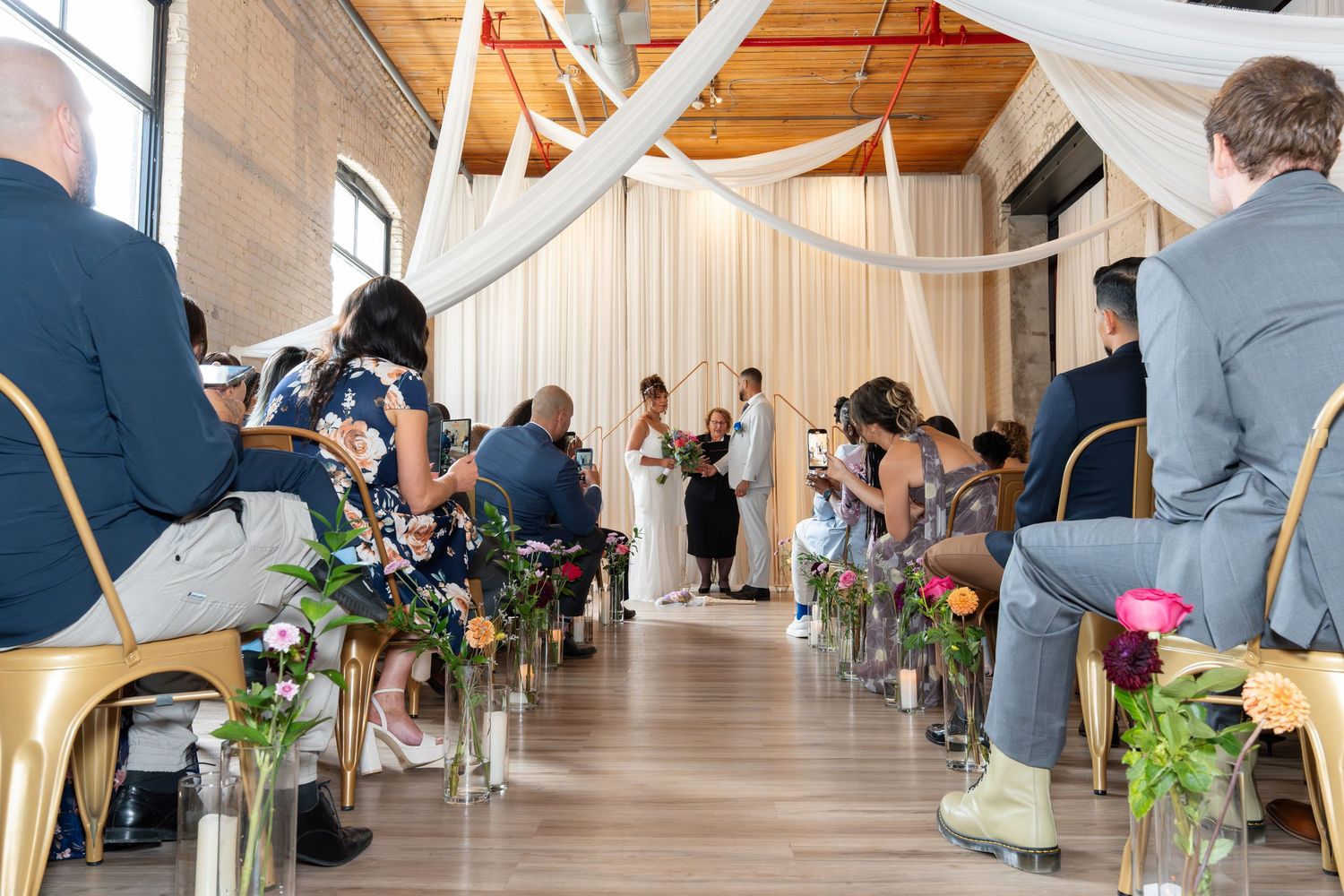 Wedding guests seated in rows of white chairs inside a bright indoor venue with white draping and floral arrangements.