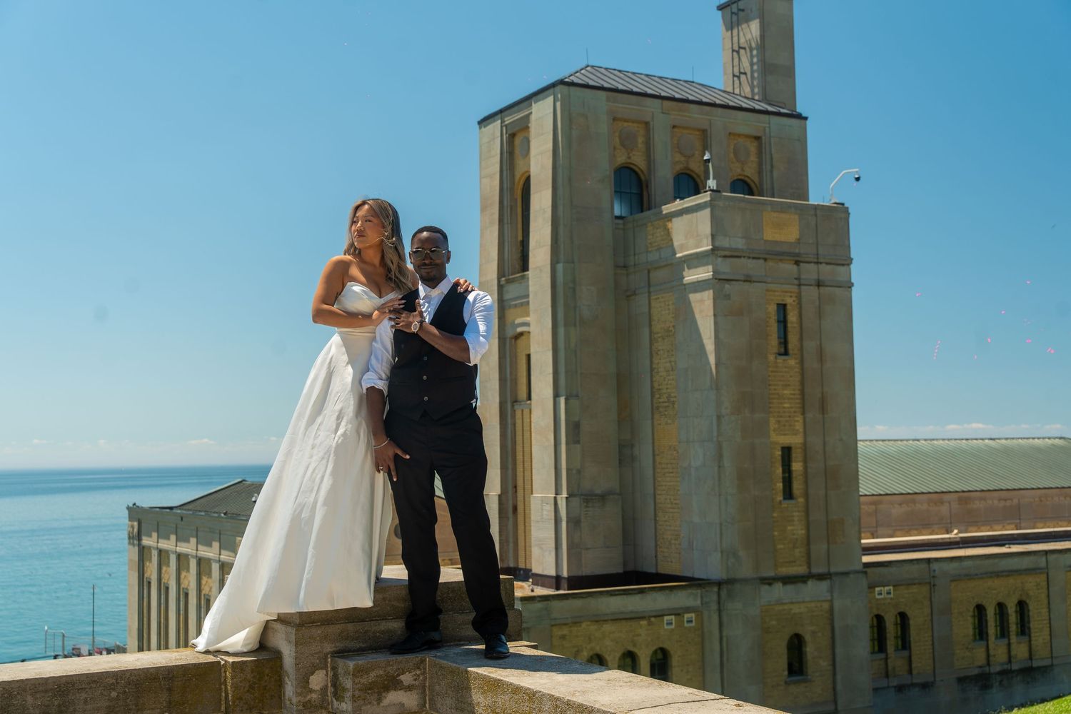 Wedding couple poses in front of stone tower buildings on a sunny day with ocean views in the background.