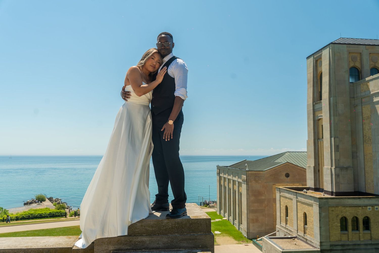 A romantic wedding couple poses by a scenic overlook against a bright blue sky.