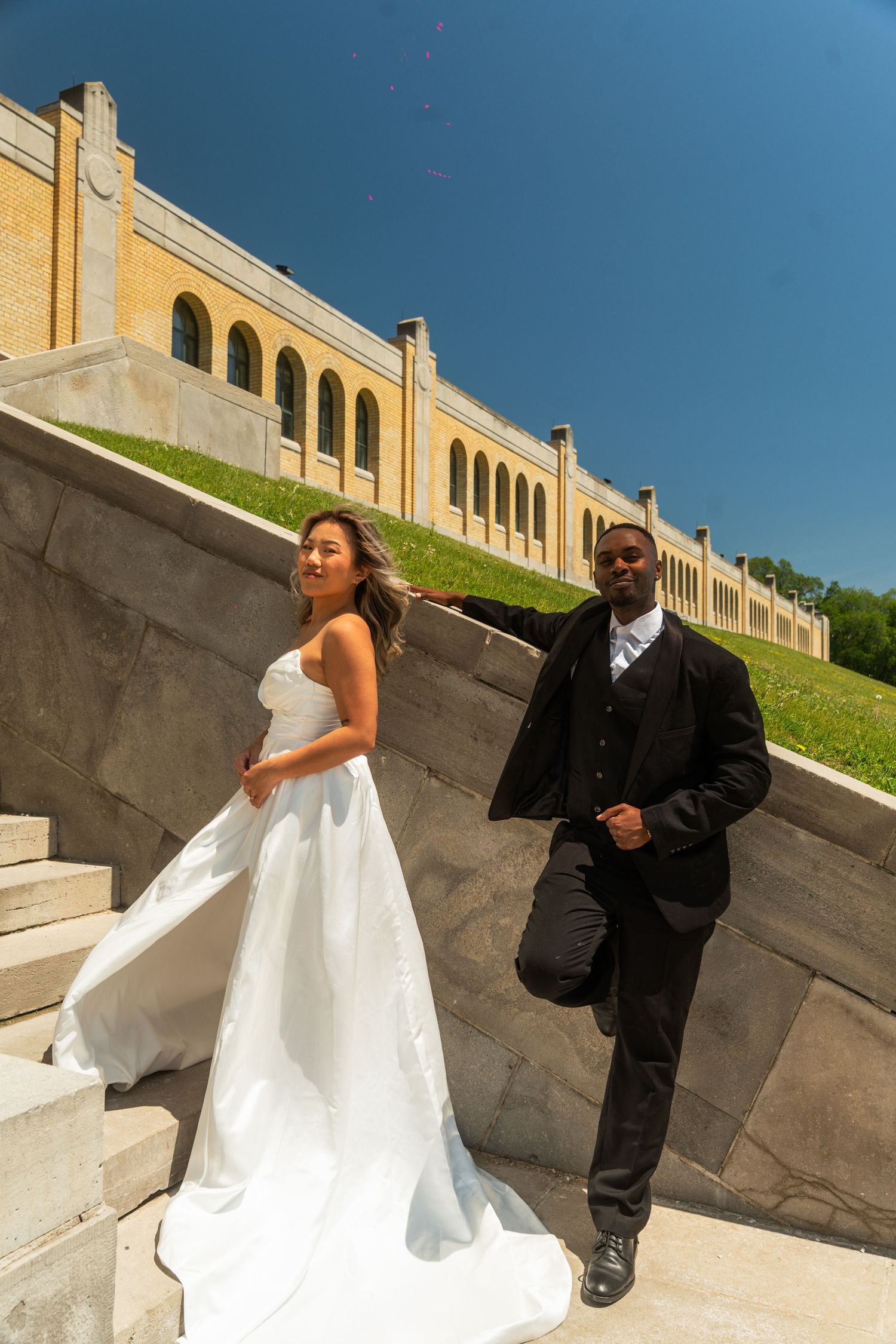 A couple in wedding attire playfully interacts on grand outdoor stone steps.