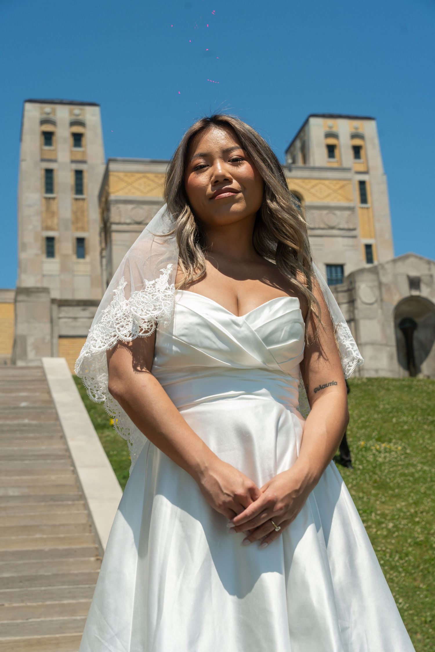 A radiant bride in a white satin gown poses on stone steps with classic architecture.