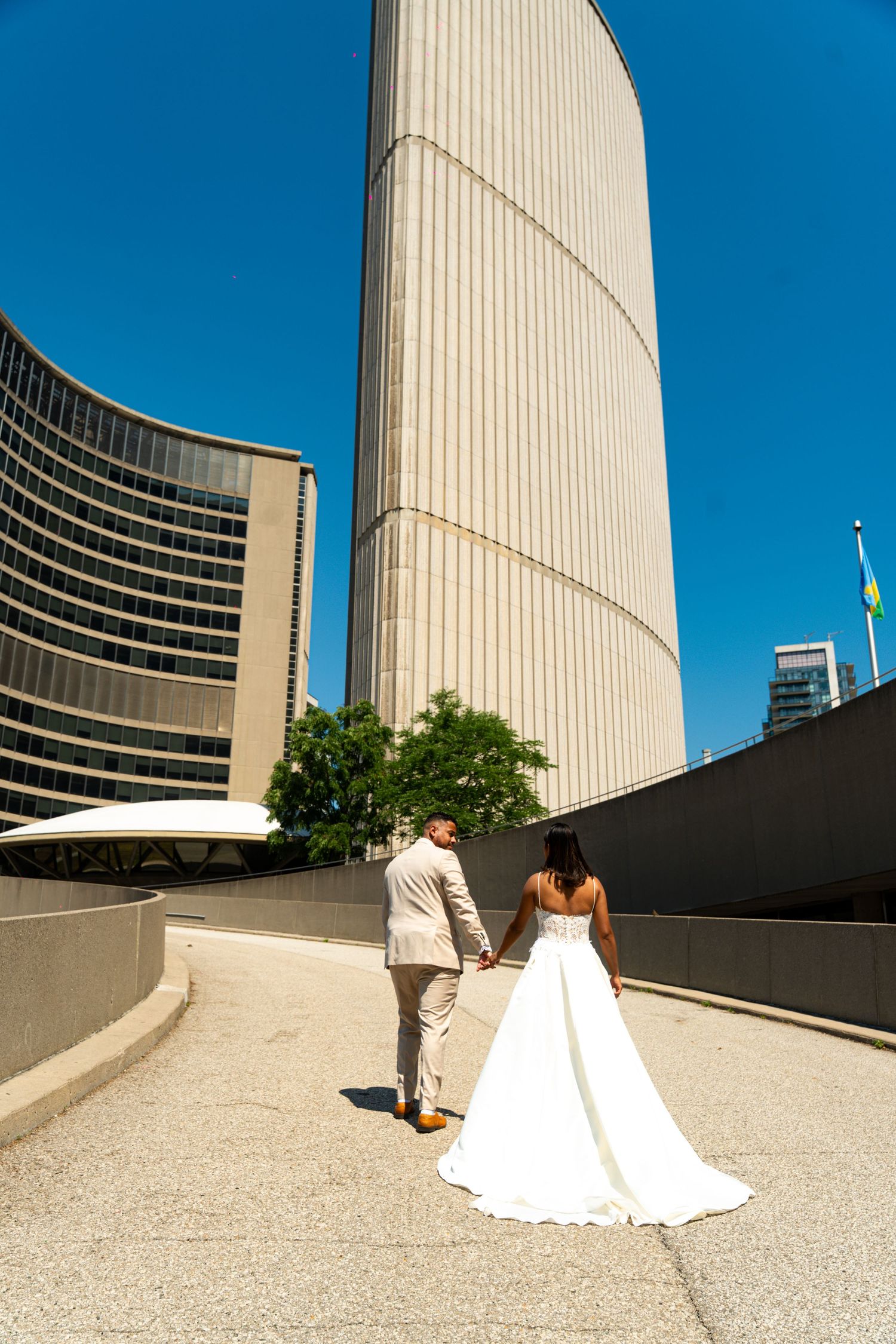Five sequential photos show a couple in wedding attire walking along a curved concrete path next to modern architecture.