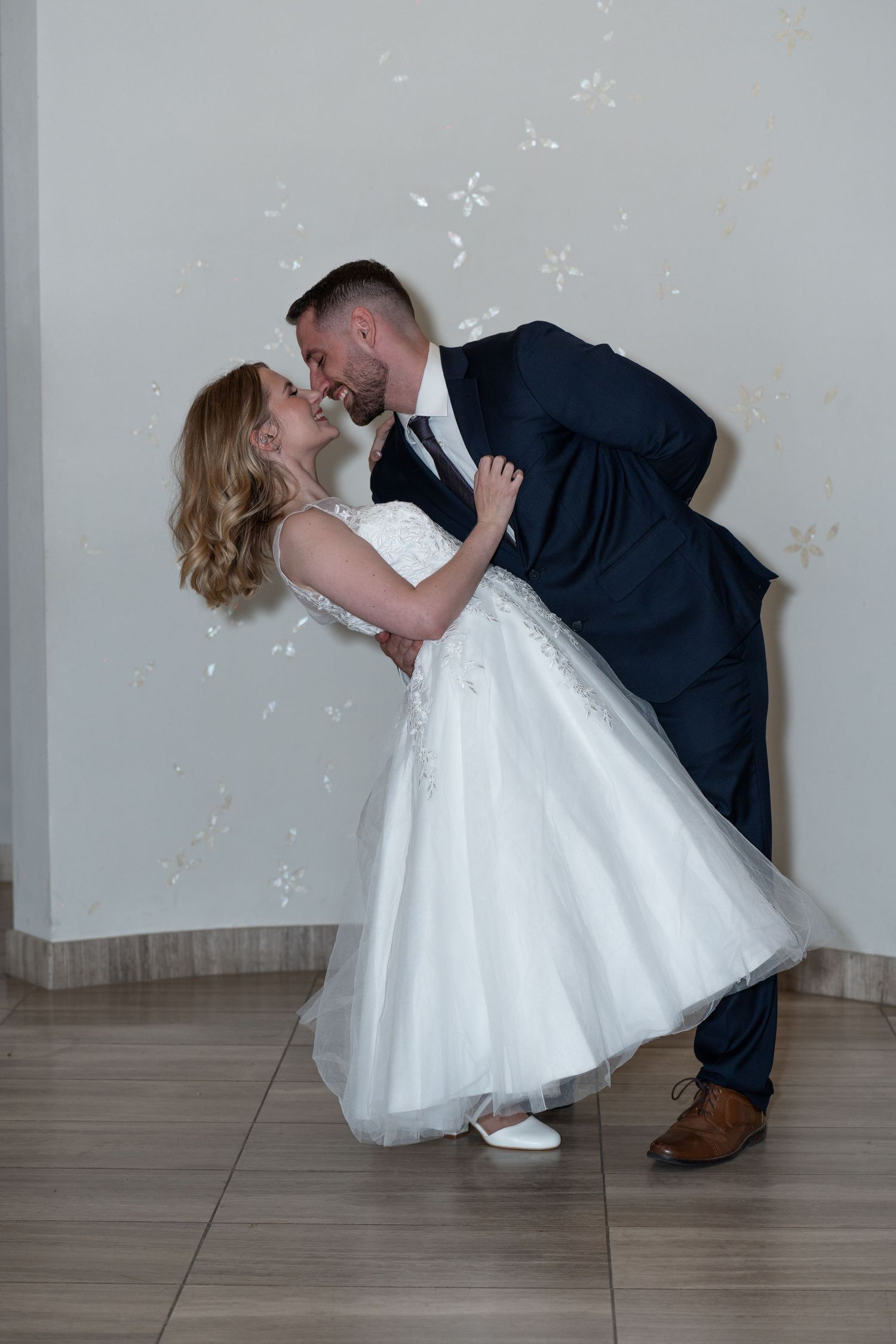 A couple in wedding attire dips and poses in a romantic dance sequence against a light gray wall.