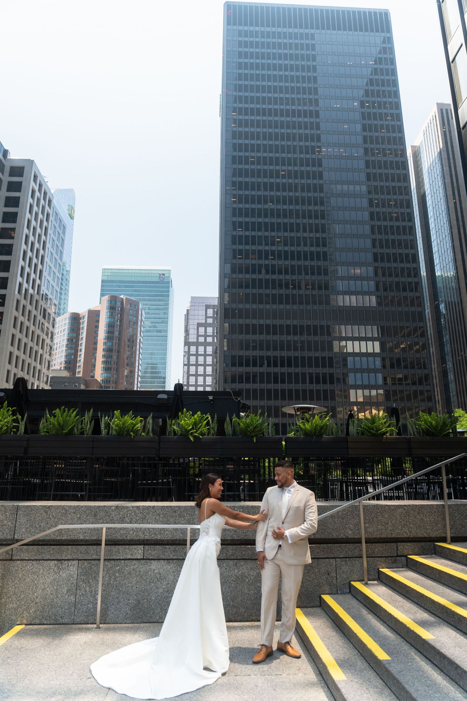 A couple in wedding attire stands against a backdrop of modern skyscrapers in an urban setting.