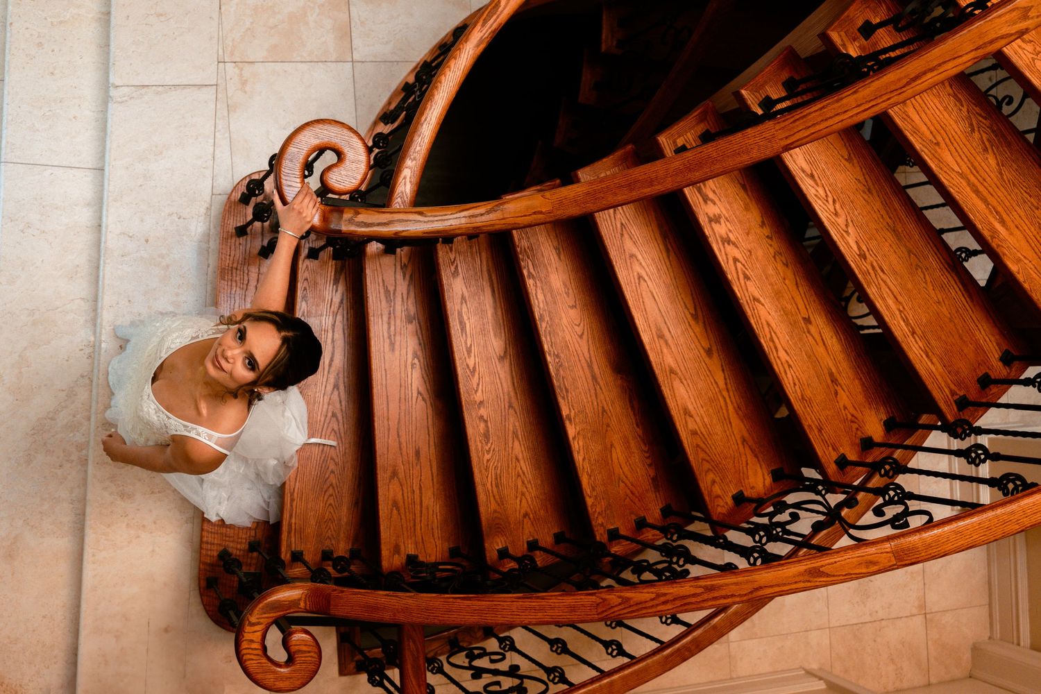 A person in white clothing ascends a curved wooden staircase with ornate railings in a dramatic architectural setting.