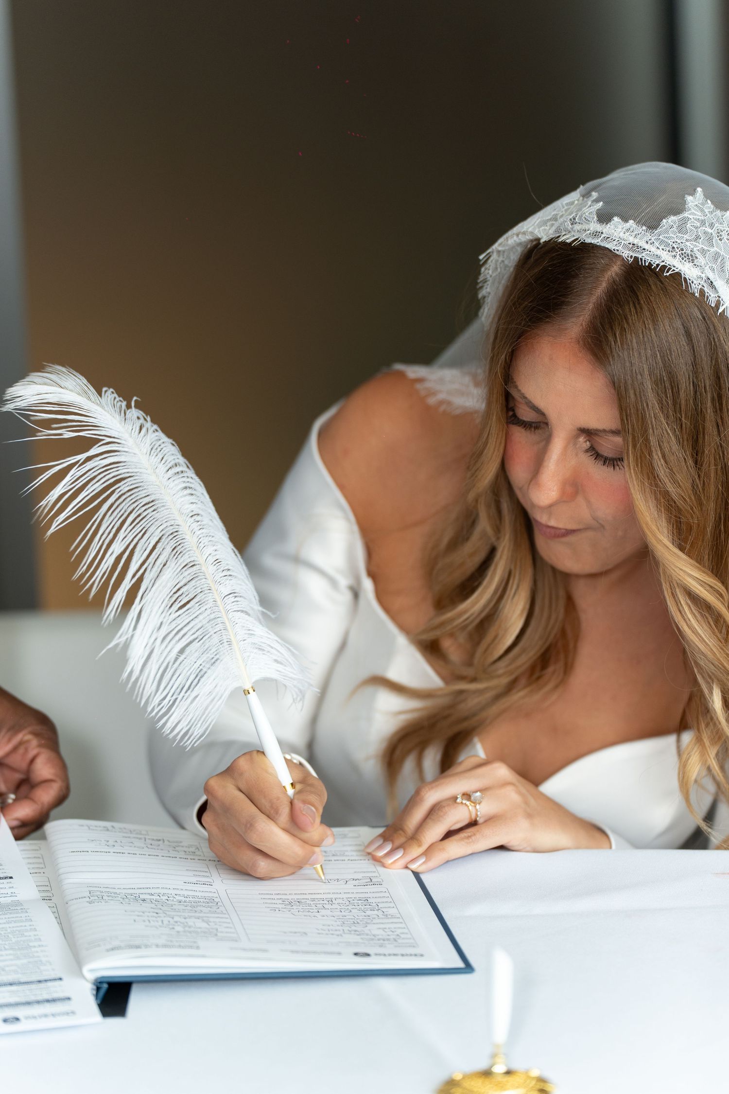 A person wearing white leans over a document to write with a white feather quill pen in an elegant setting.