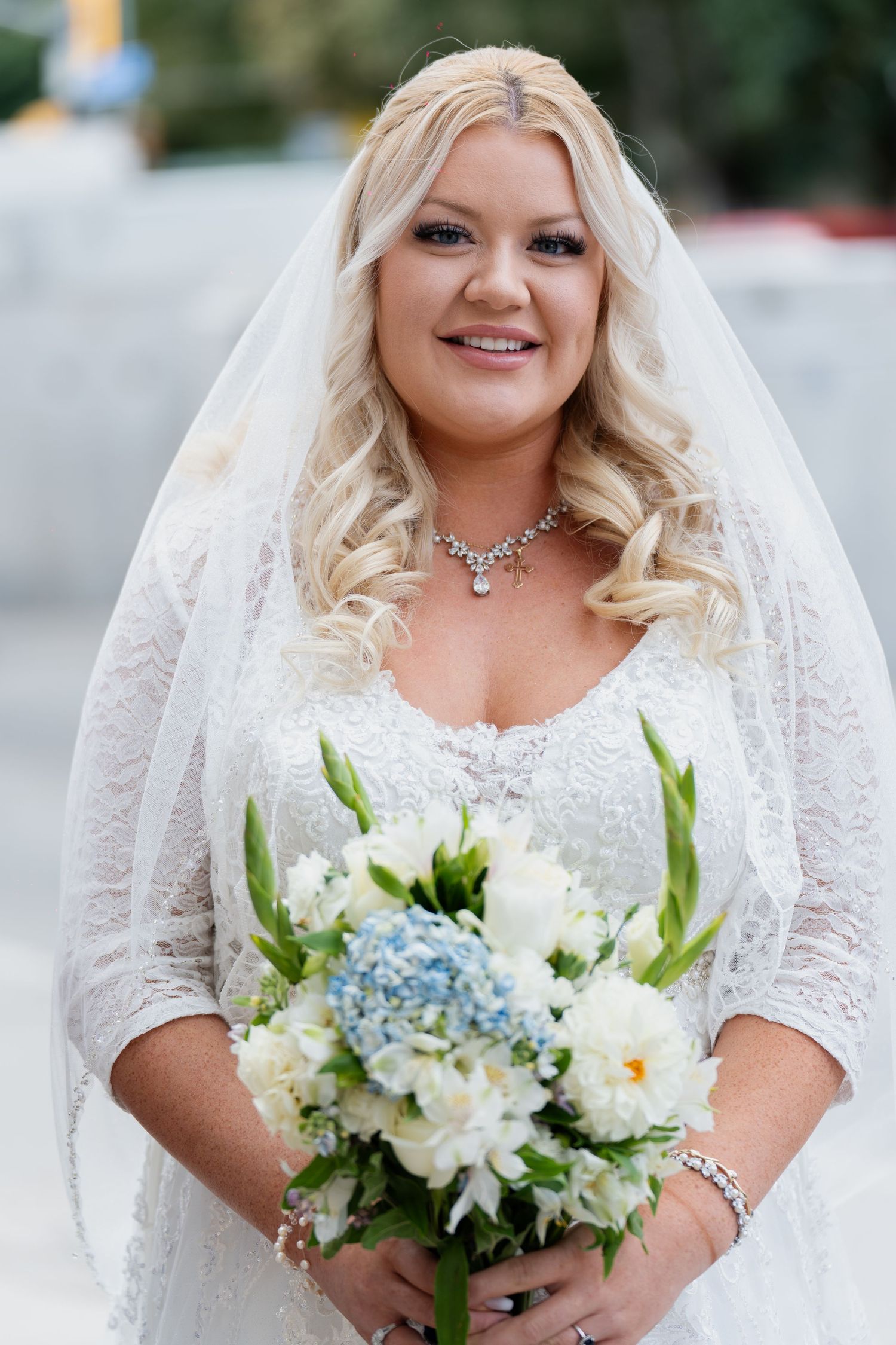 Bride in white lace dress holds elegant bouquet of blue hydrangeas and white flowers with greenery accents.