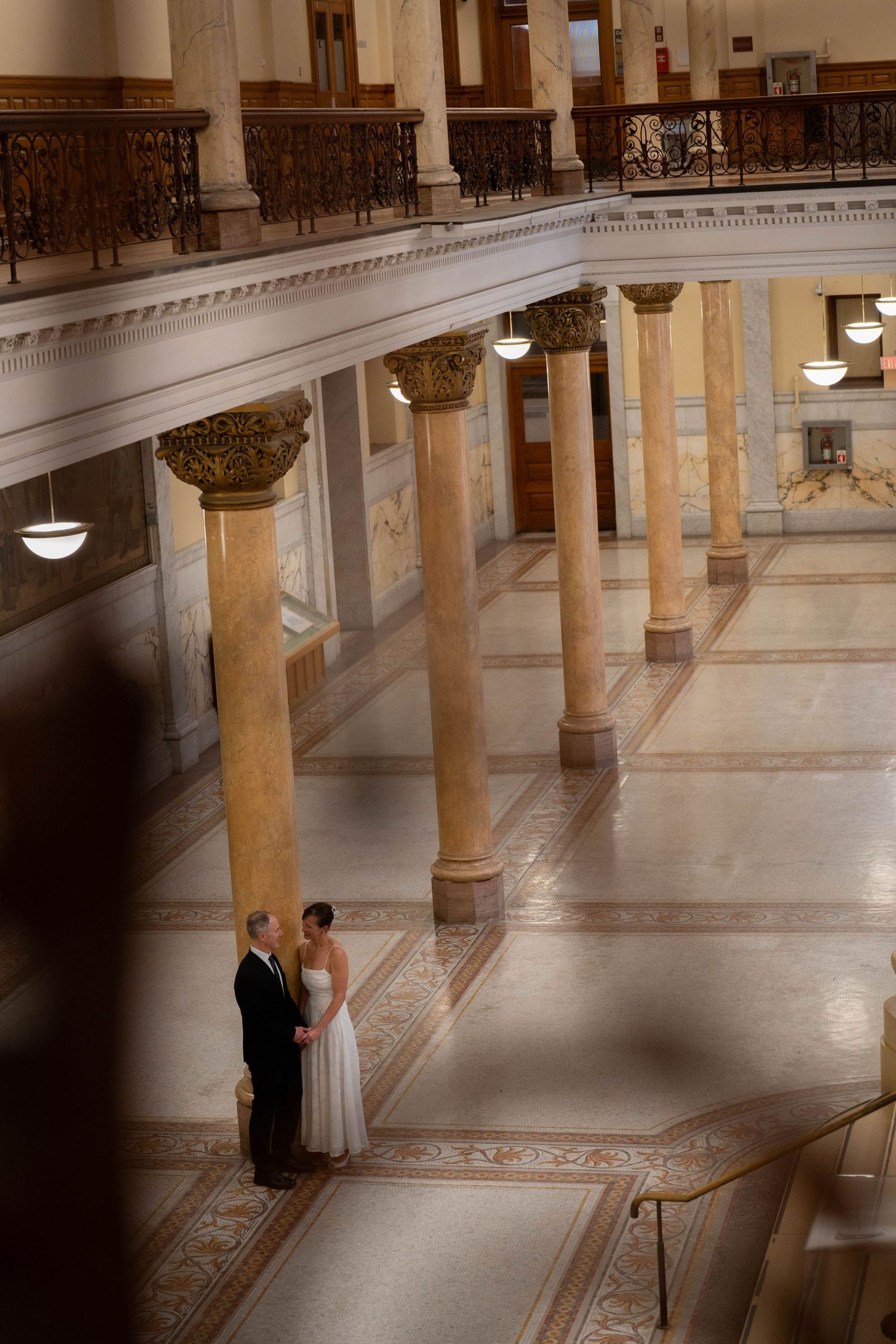 Bride and groom stand together in grand marble hall with tall columns and ornate balconies during wedding ceremony.