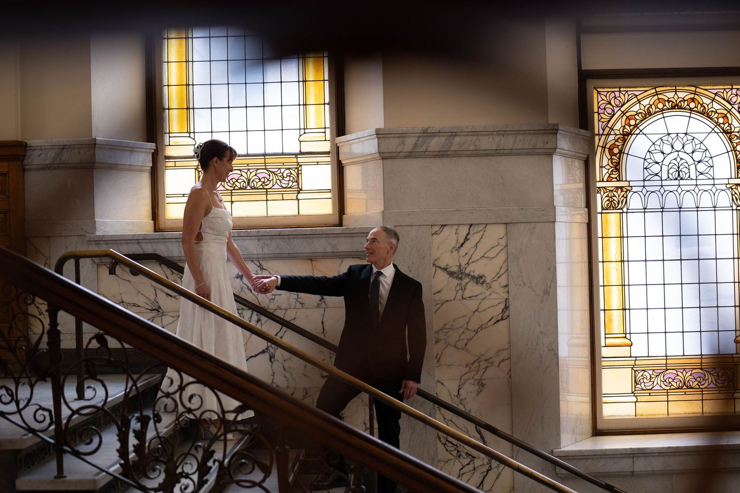 Bride and groom hold hands on grand marble staircase with ornate stained glass windows in elegant wedding venue.