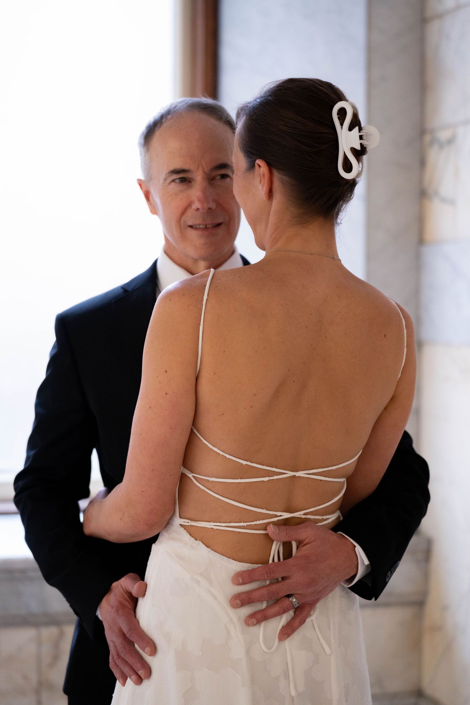Couple sharing a tender moment, bride in elegant backless white gown with criss-cross straps and groom in dark suit.