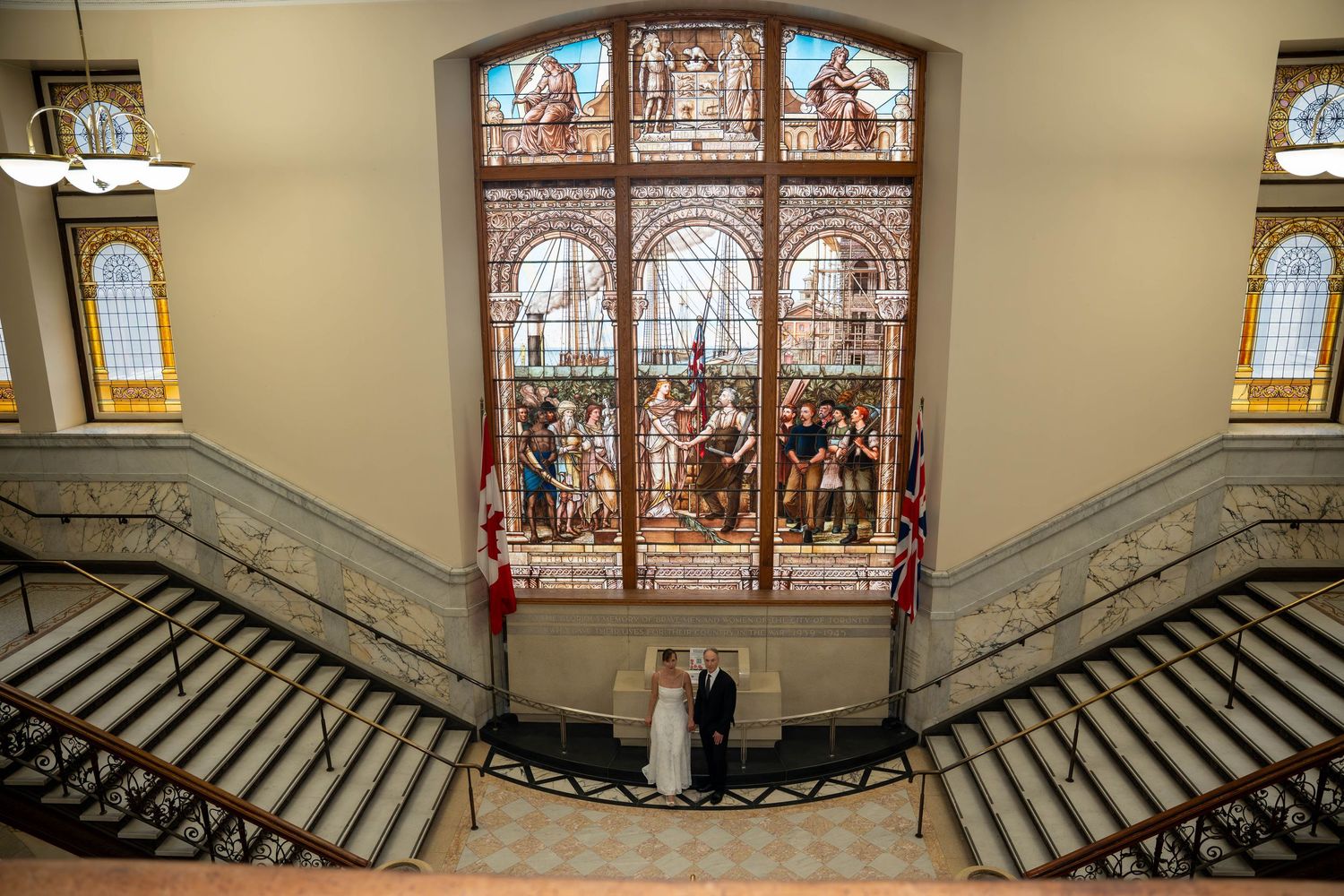 Bride and groom pose on grand staircase beneath ornate stained glass window in elegant historic building.