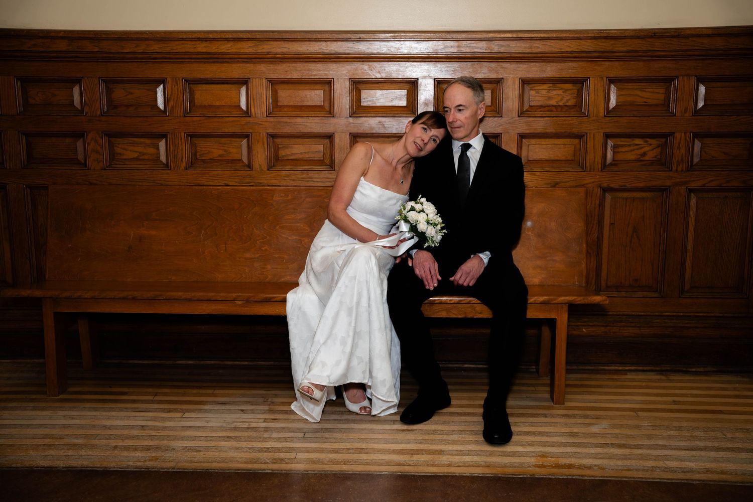 Bride and groom sit closely together on a wooden bench, bride holding a white bouquet.
