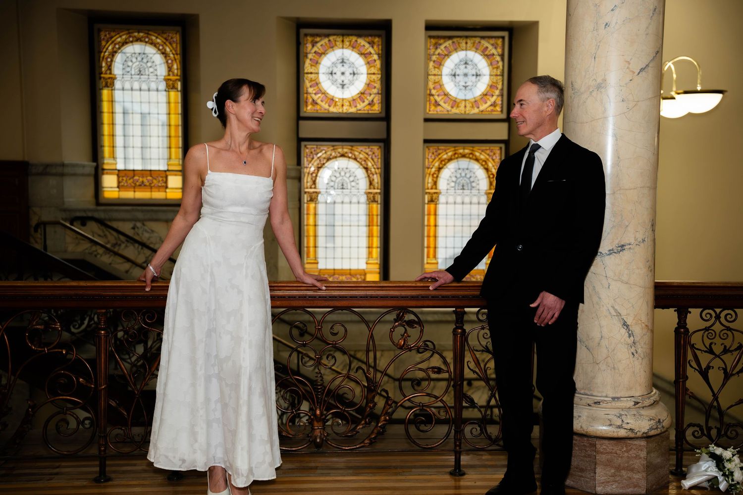 Bride and groom share a romantic moment on an ornate staircase with stained glass windows.