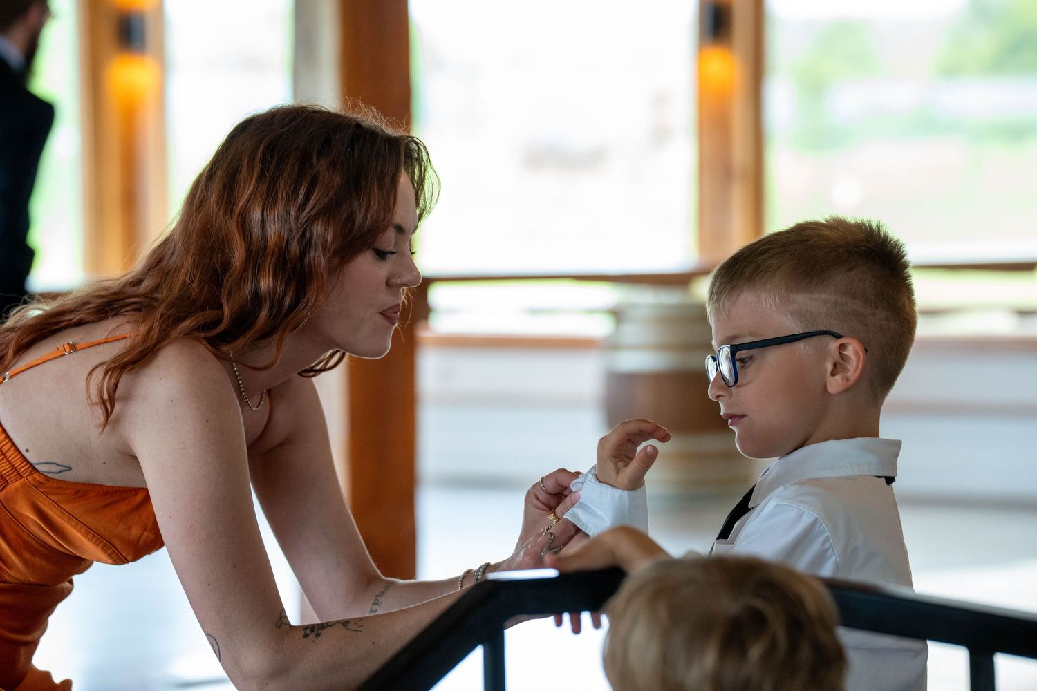 Woman in orange dress bending down to talk to young boy in glasses and white shirt at a formal event.