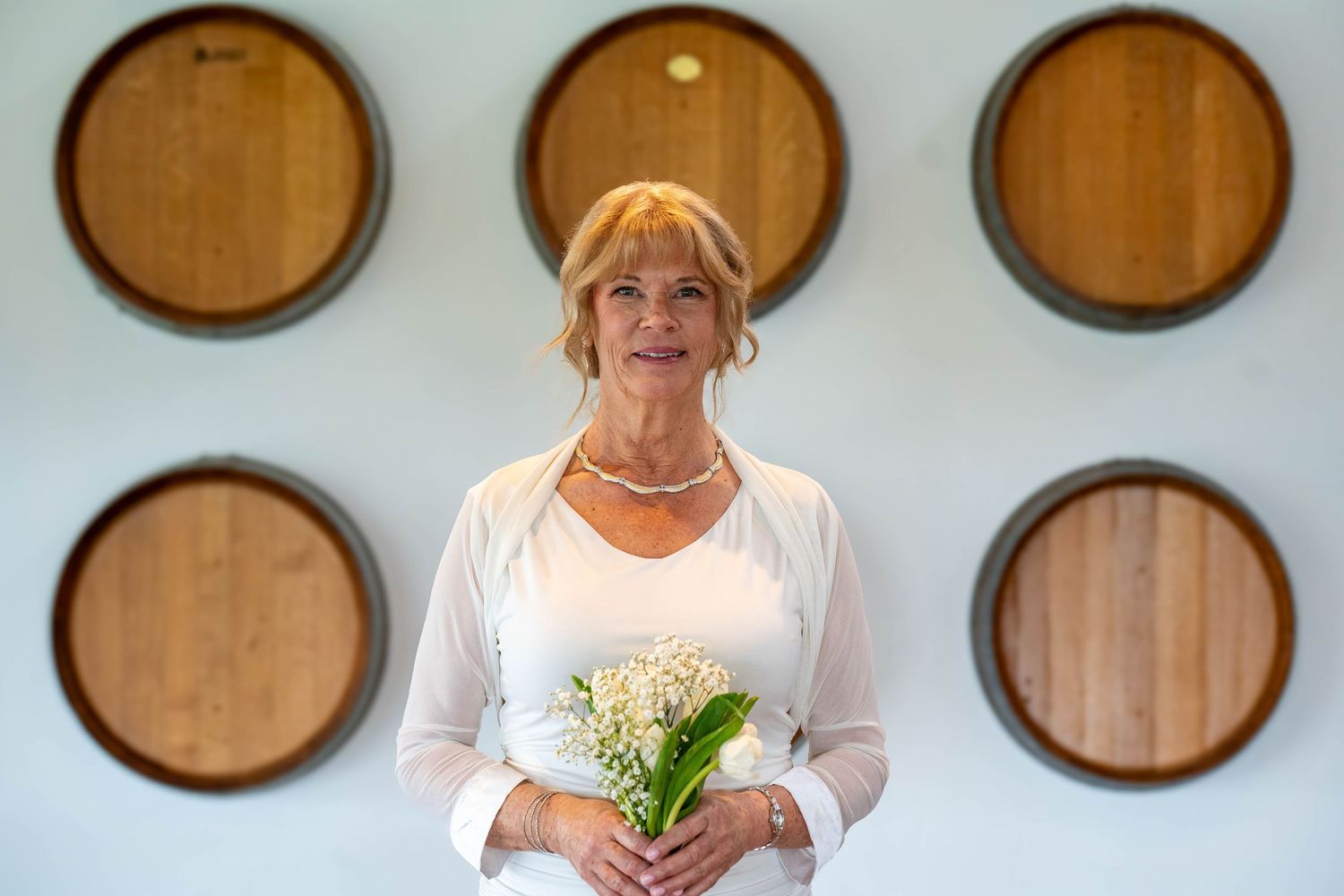 Smiling blonde woman in white outfit holding lily of the valley flowers, standing before decorative wooden barrel wall art.