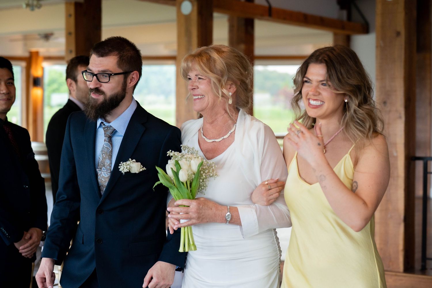 Smiling wedding guests in formal attire, including a woman in white holding white flowers, stand together at an indoor venue.