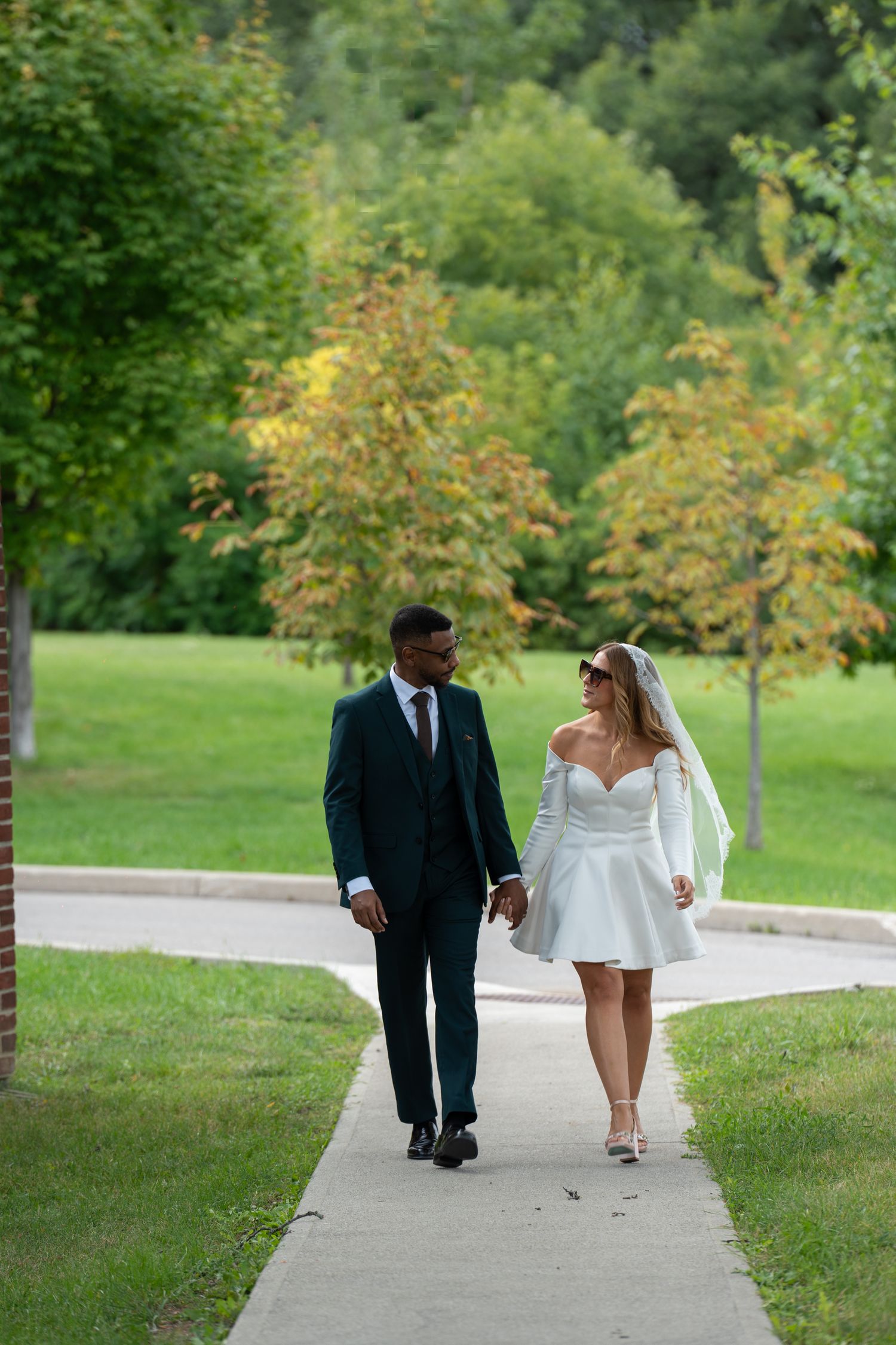 A couple walks together on a winding concrete path through a lush green park setting on a sunny day.