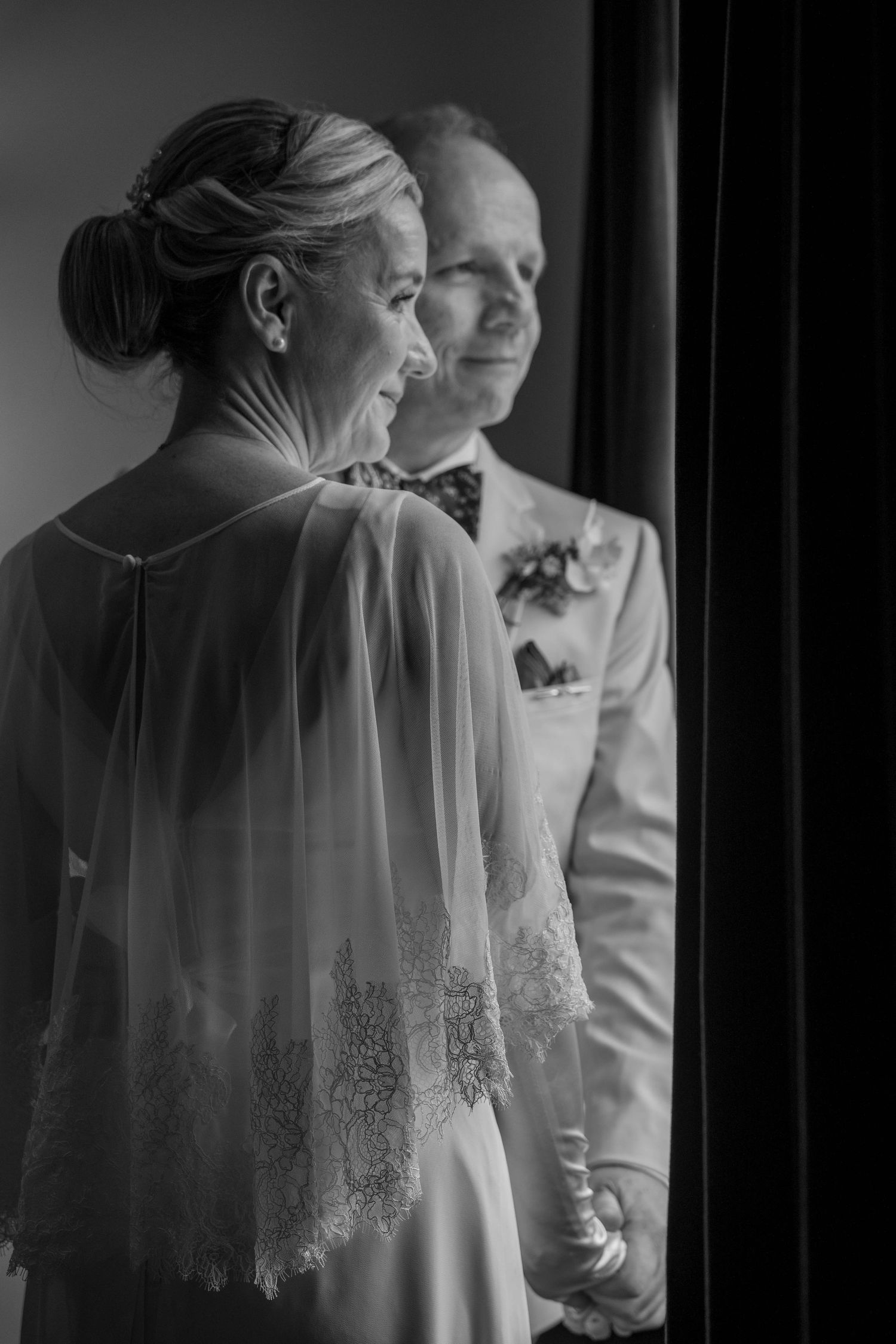 Black and white photo of an elegant bride in lace cape gown standing with a smiling groom in a suit at their wedding.