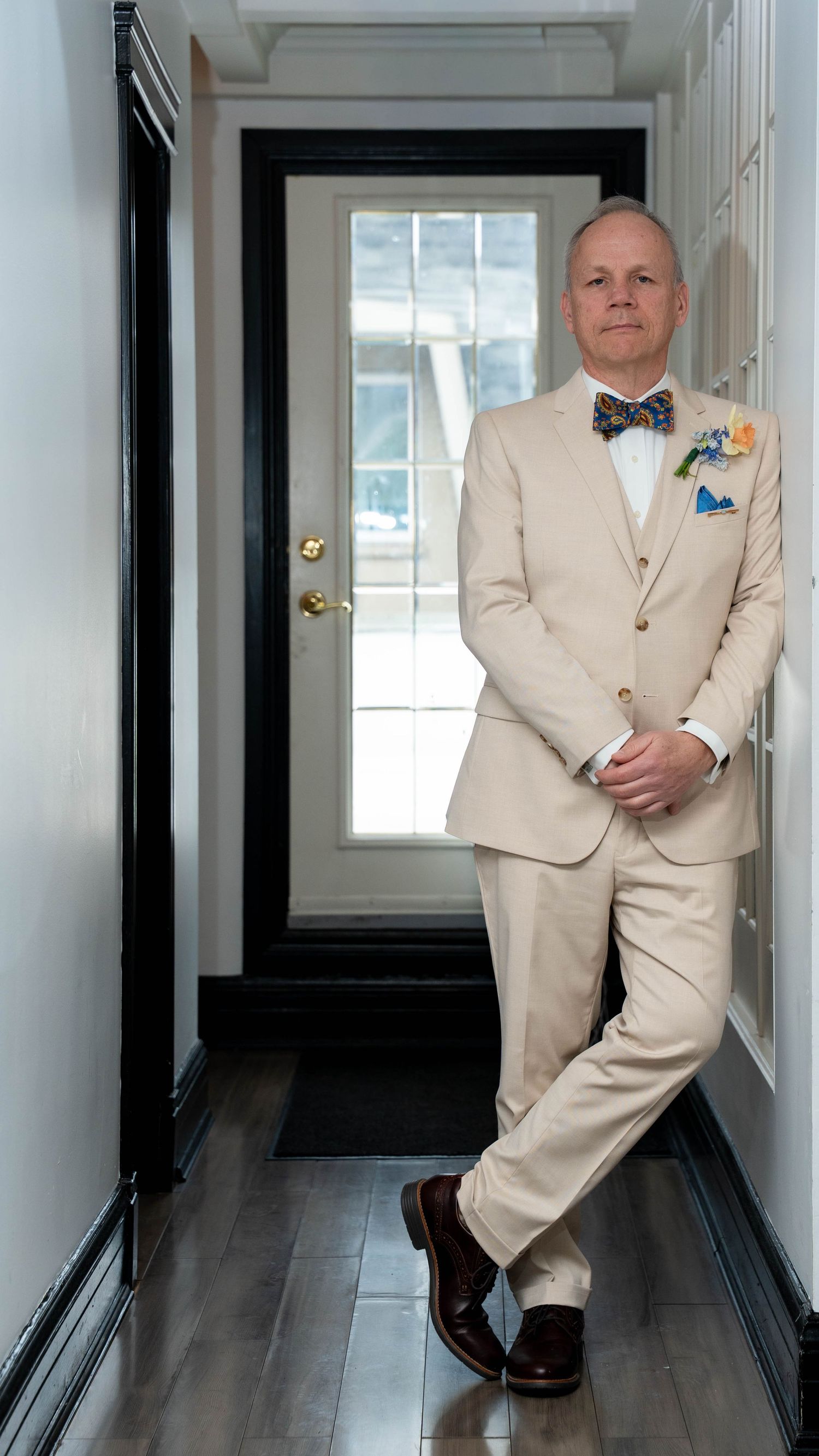 Man in beige suit with colorful bow tie and boutonniere stands confidently in a hallway with dark hardwood floors.