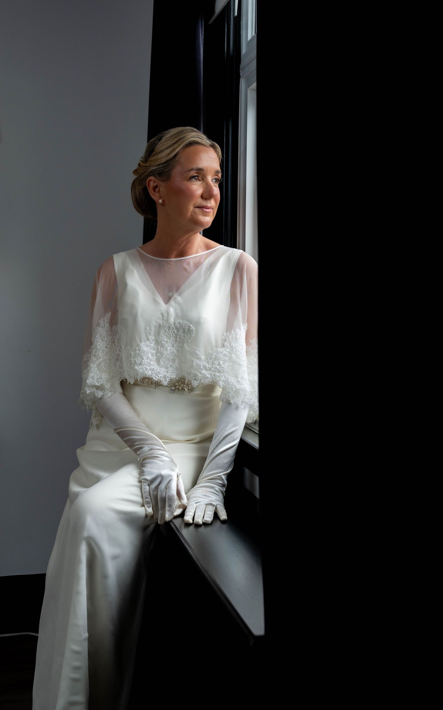 Elegant bride in white lace capelet, satin pants, and long gloves sits by a window gazing thoughtfully into the distance.