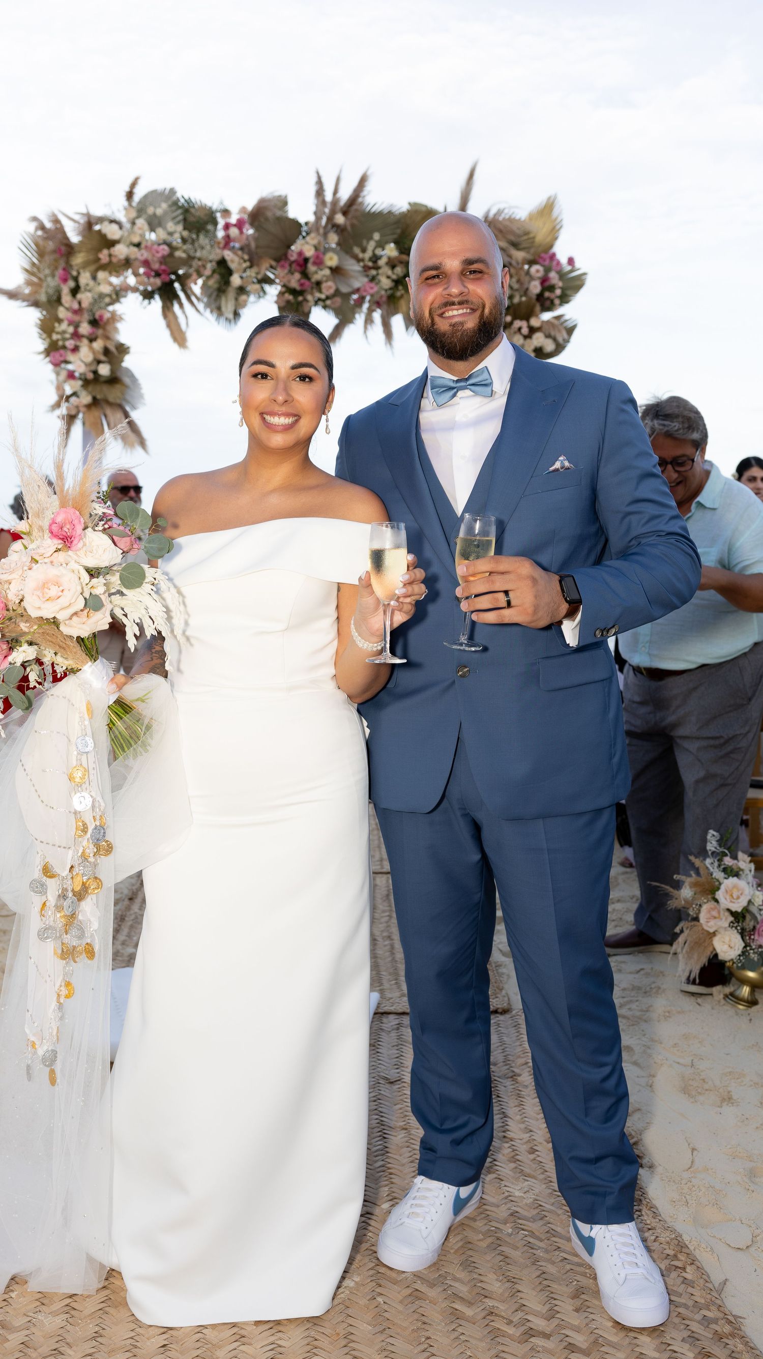 Beach wedding celebration with couple toasting under floral arch with tropical decor.