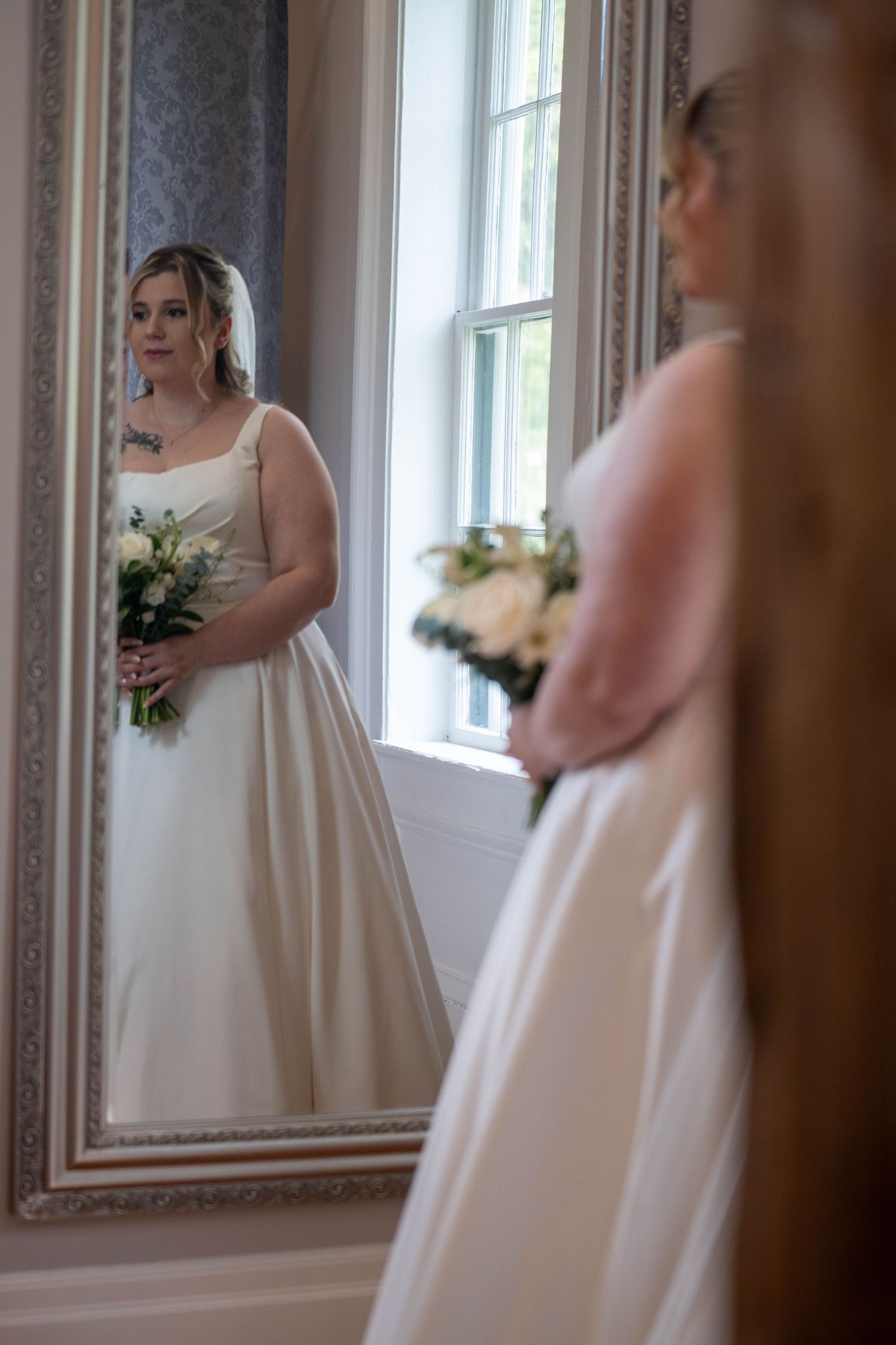Bride in white gown holding bouquet reflected in ornate mirror near window.