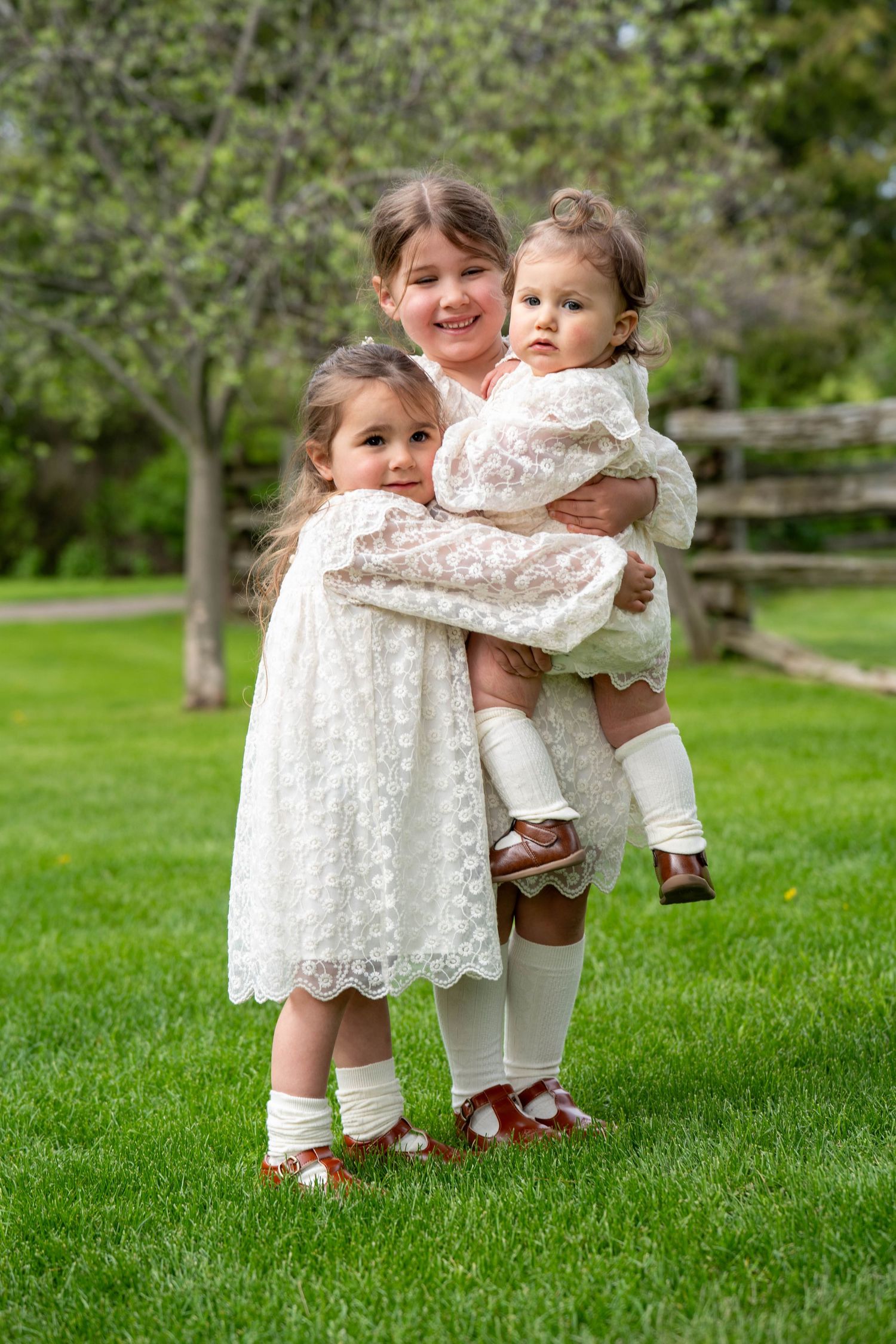 Three young girls in white lace dresses pose together on green grass, with the oldest holding the youngest.