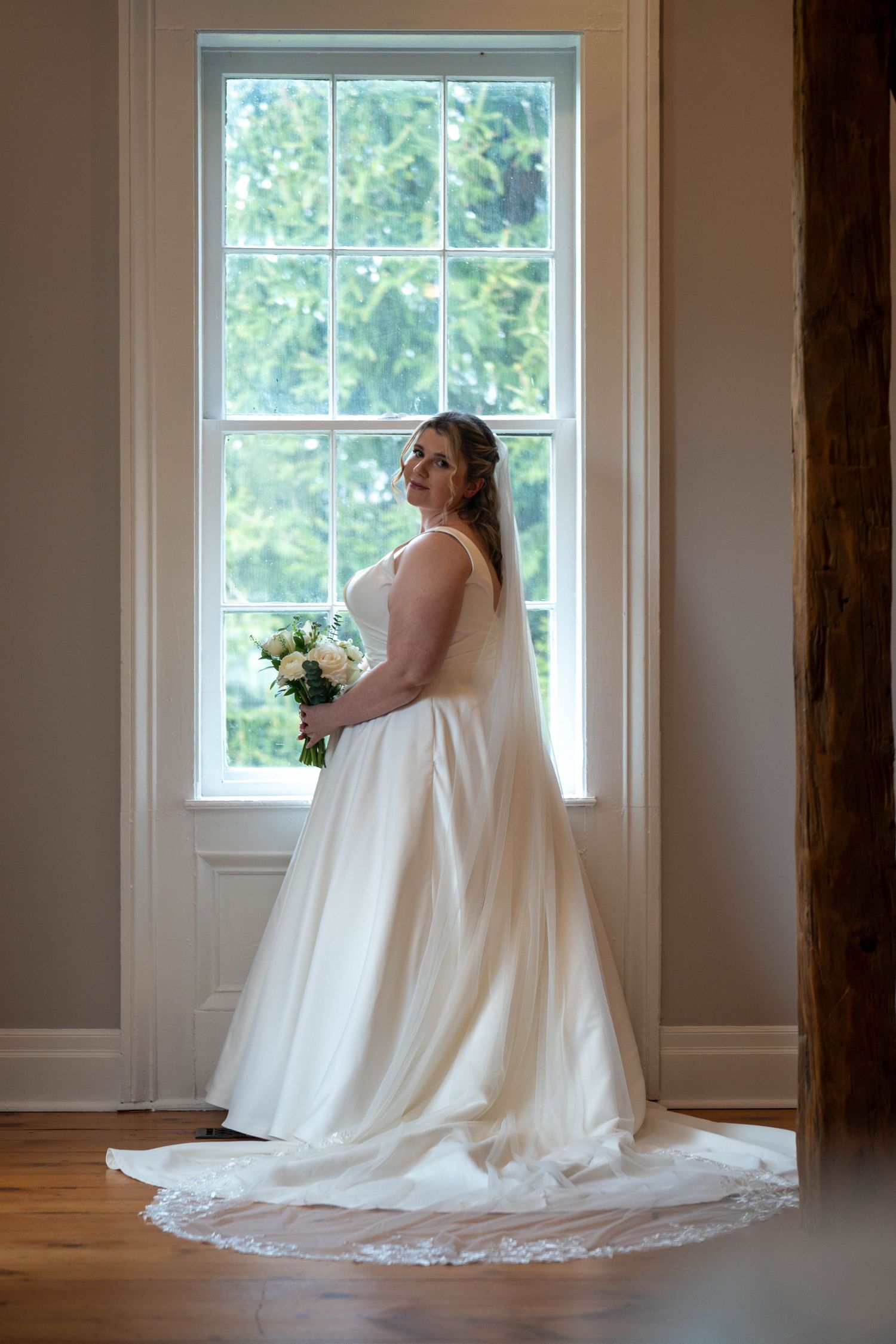 Bride in elegant white gown with train holding bouquet standing by sunlit window.