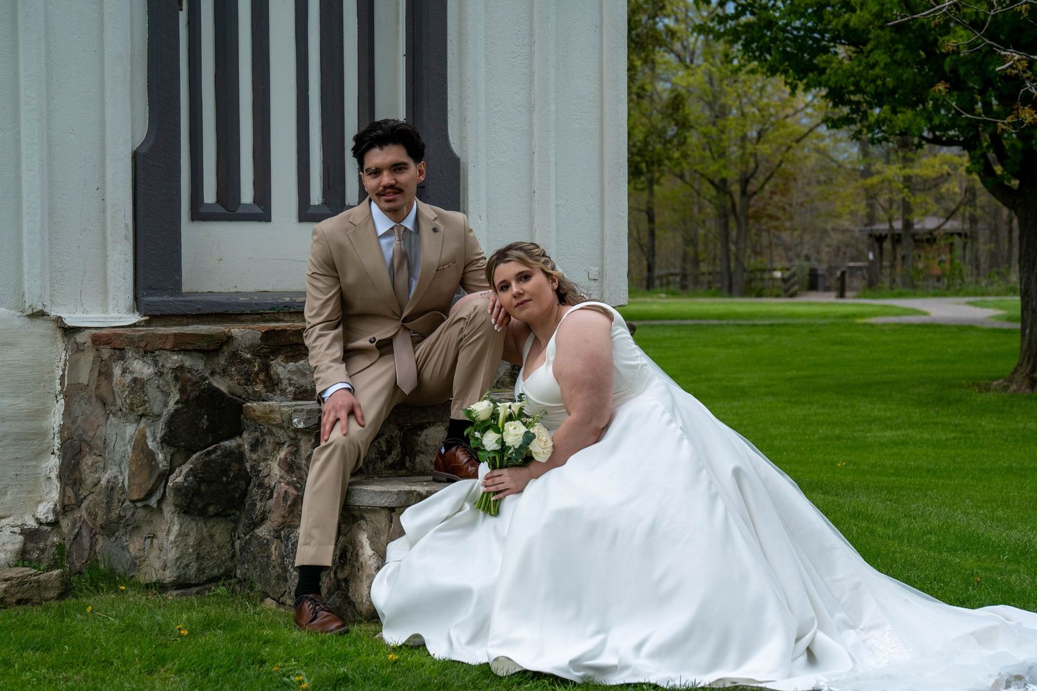 Bride in white gown and groom in tan suit pose on stone steps outside a white building on their wedding day.