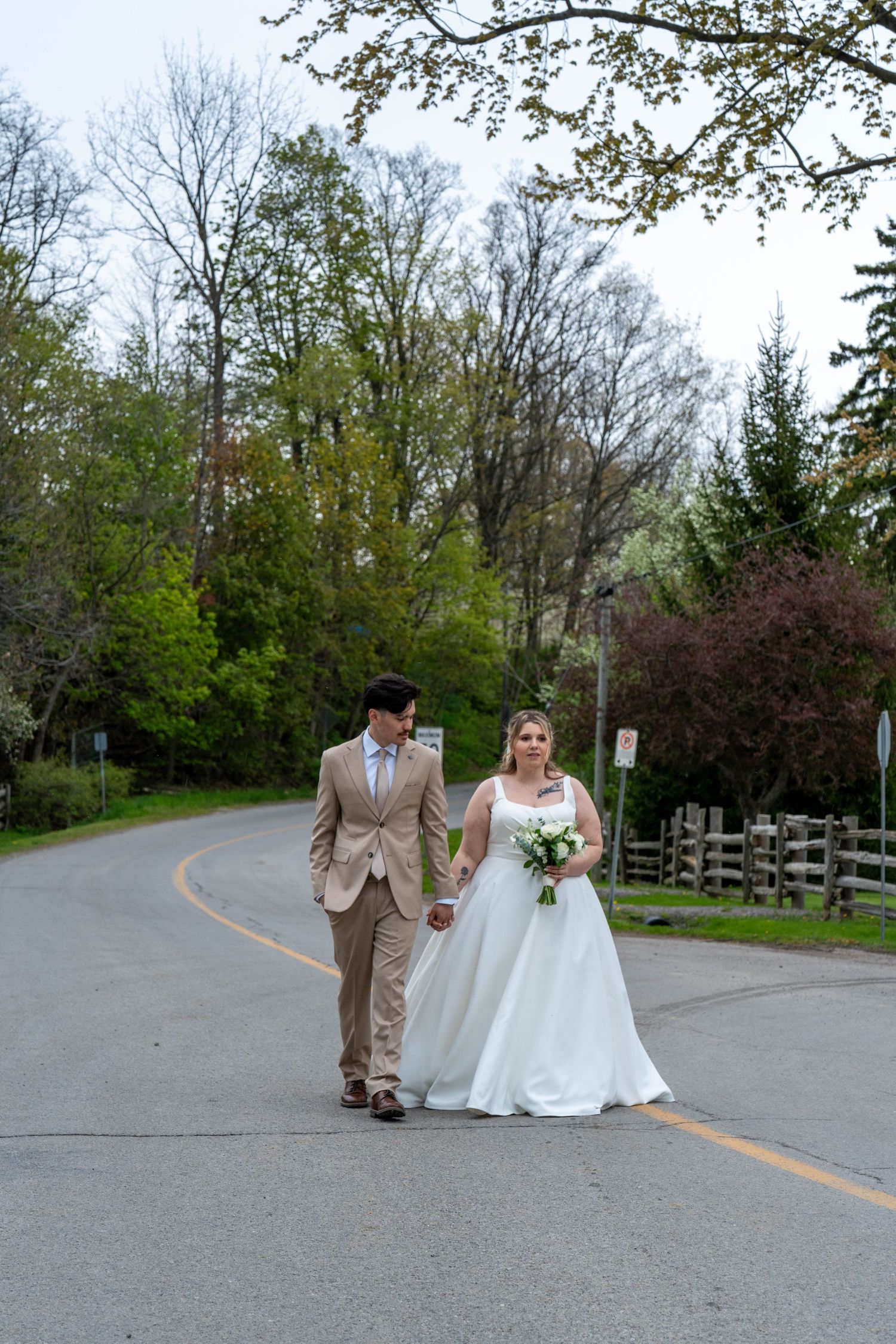 Bride and groom walking hand in hand on a road surrounded by lush green trees on their wedding day.