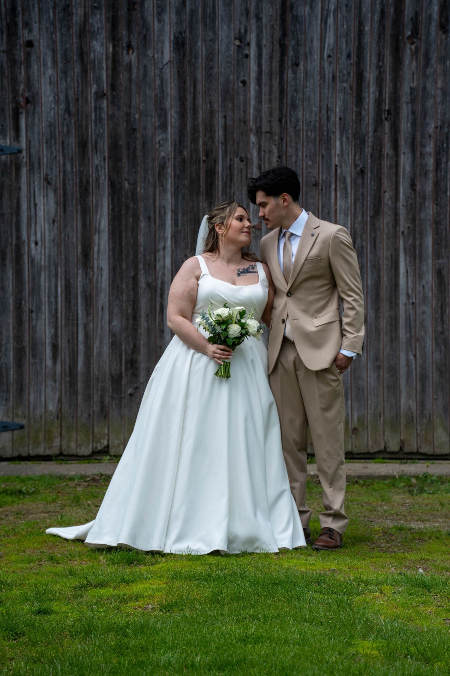 Bride in white gown holding bouquet stands with groom in tan suit before rustic wooden barn wall on green grass.